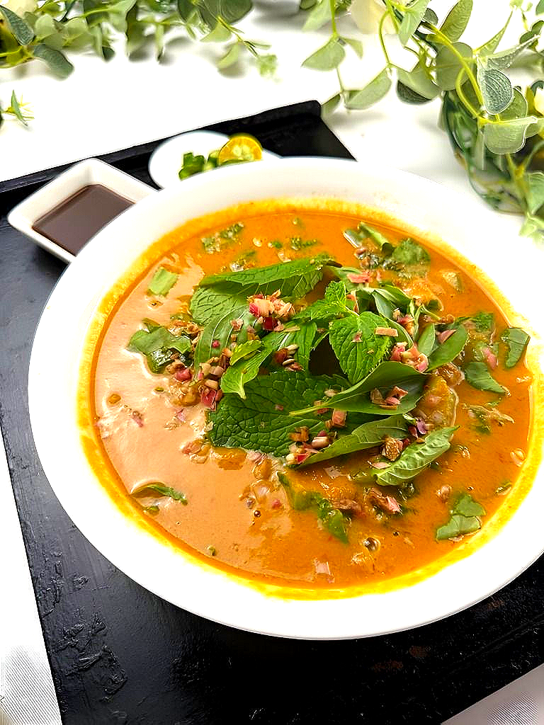 Bowl of Crab Laksa, topped with herbs, next to small dipping sauce and garnish on a black tray.