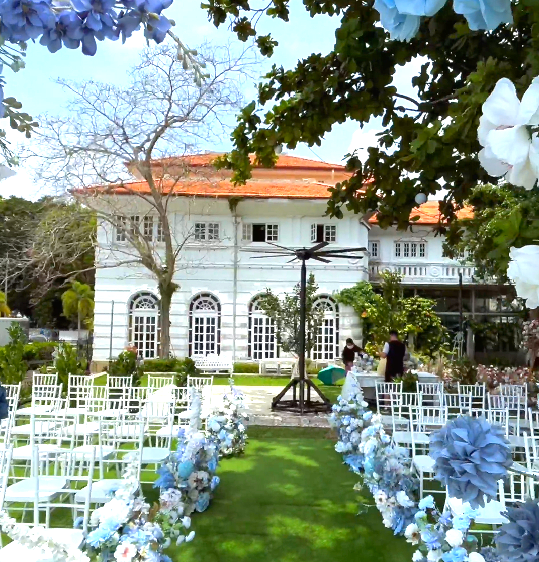 A white mansion with a tiled roof sits behind an outdoor wedding aisle lined with white chairs and blue floral displays.