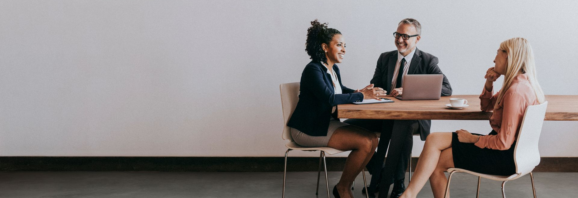 Three people in business attire at a table with laptop, smiling and talking in a modern room.