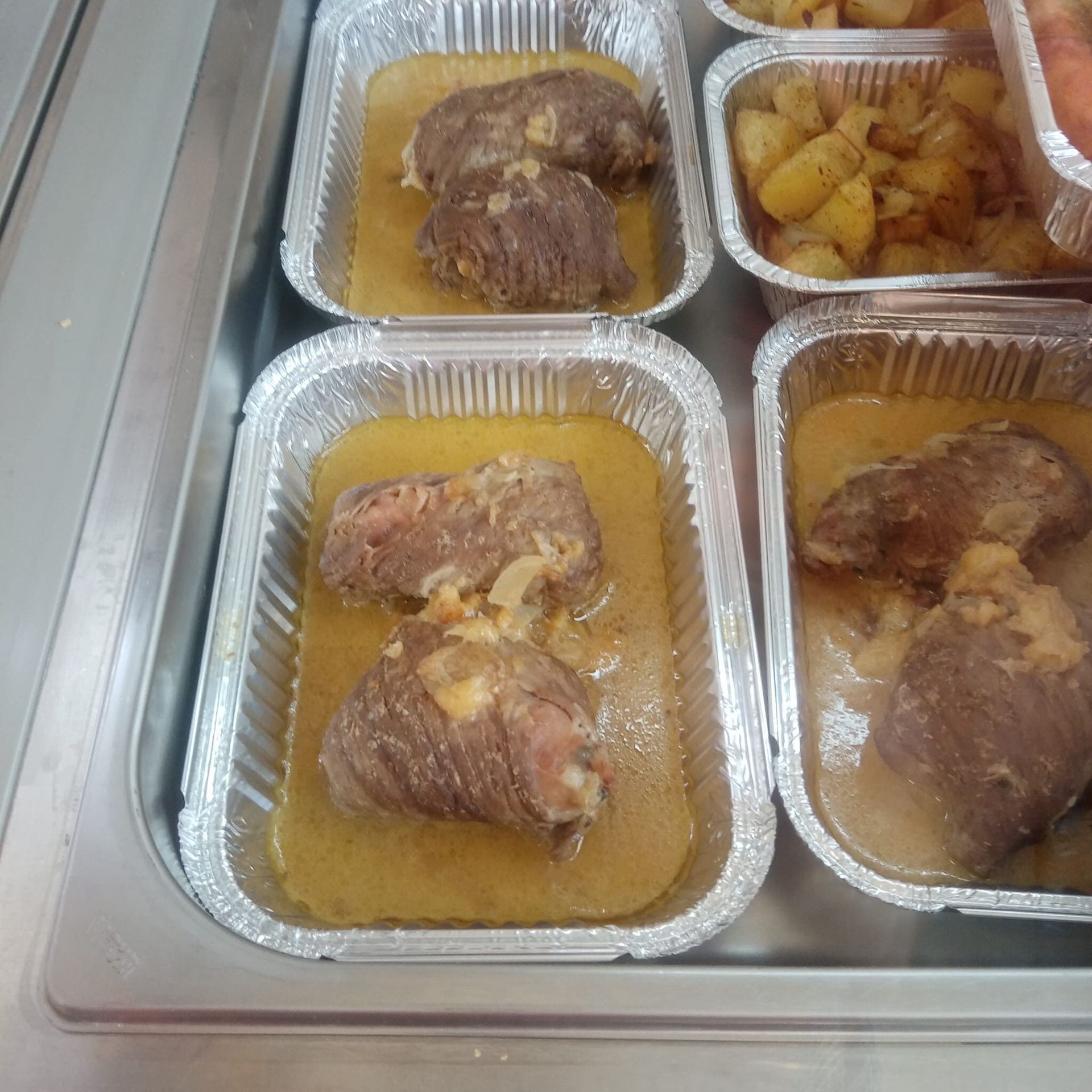 Vassoi di alluminio pieni di carne arrotolata nel sugo, patate e altri piatti disposti su una superficie metallica.