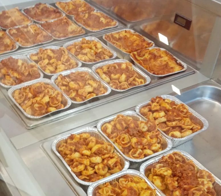 Piatti di pasta preparati in contenitori di alluminio esposti in un banco gastronomia refrigerato.