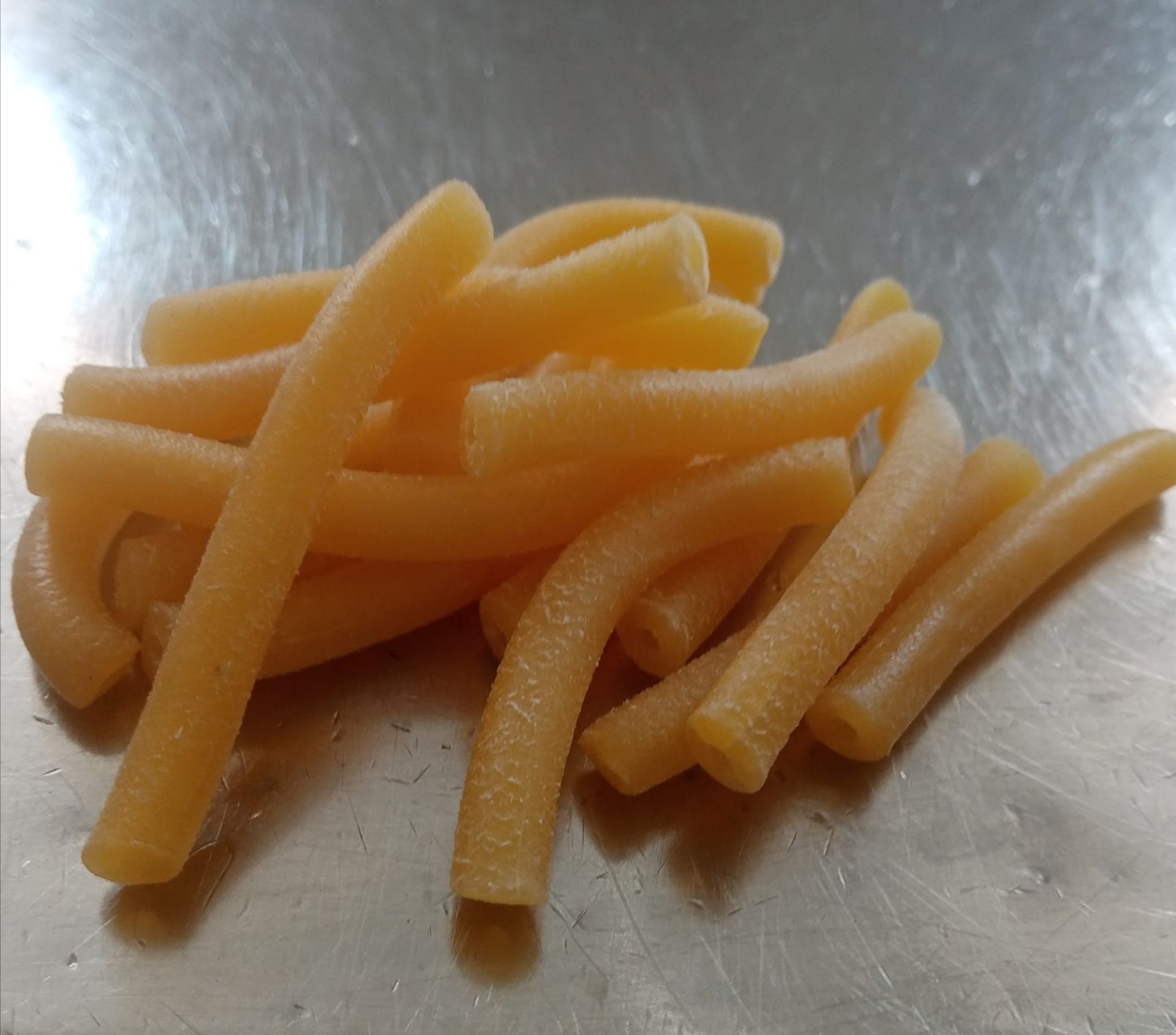 Mucchio di pasta cruda, di colore giallo dorato, su una superficie metallica riflettente.