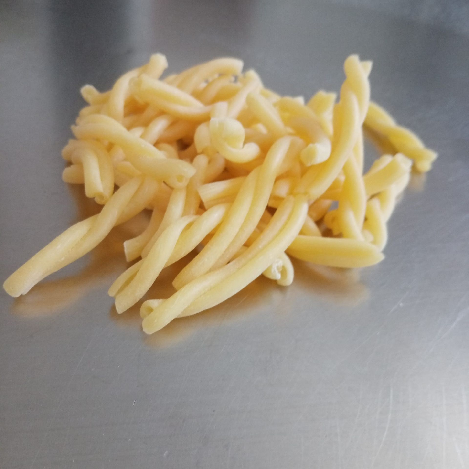 Mucchio di pasta cavatelli cruda, di colore giallo chiaro, su una superficie metallica lucida.