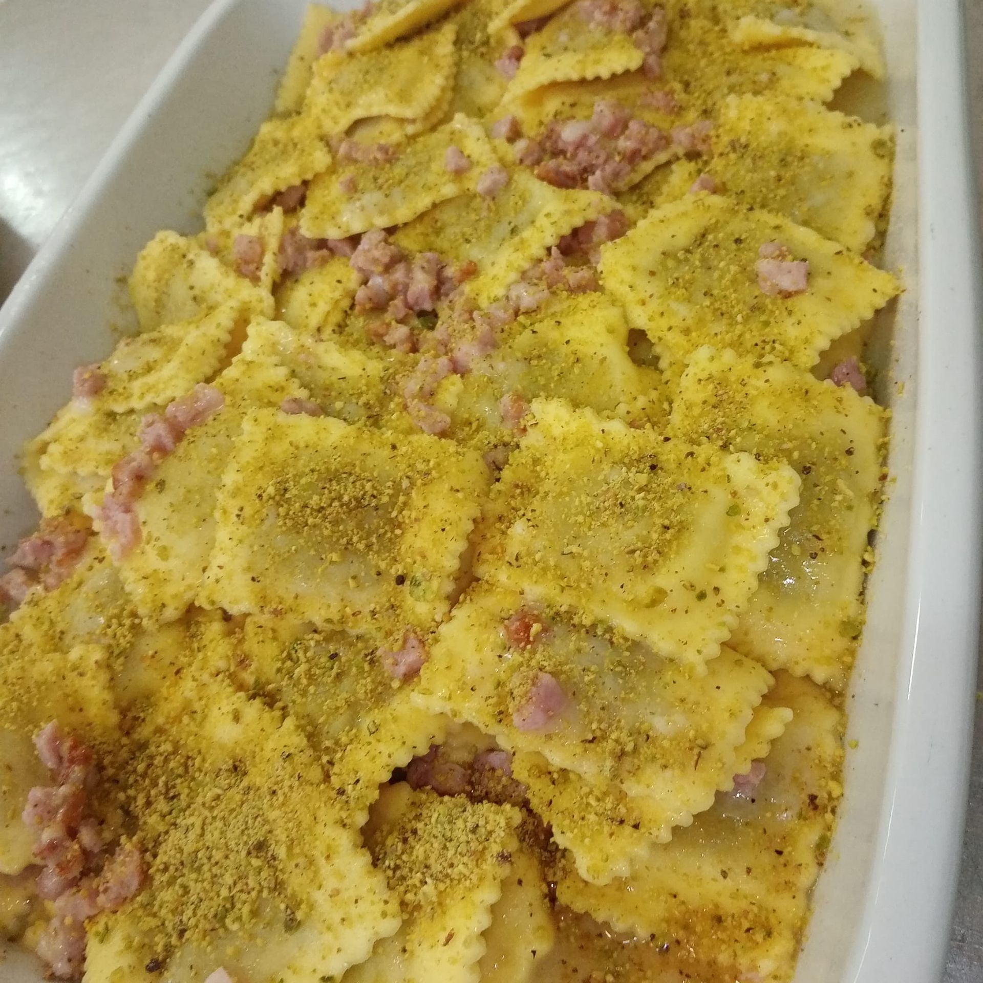 Piatto di pasta ravioli, gialli, ricoperti di carne rosa e polvere verde, in una teglia rettangolare bianca.