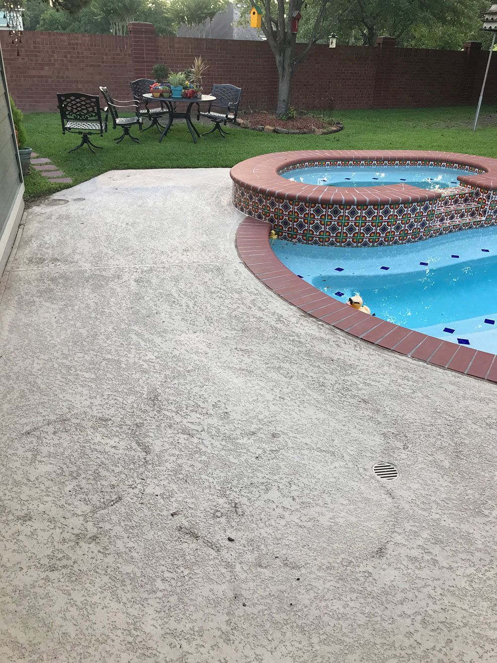 There is a swimming pool in the backyard with a table and chairs in the background.