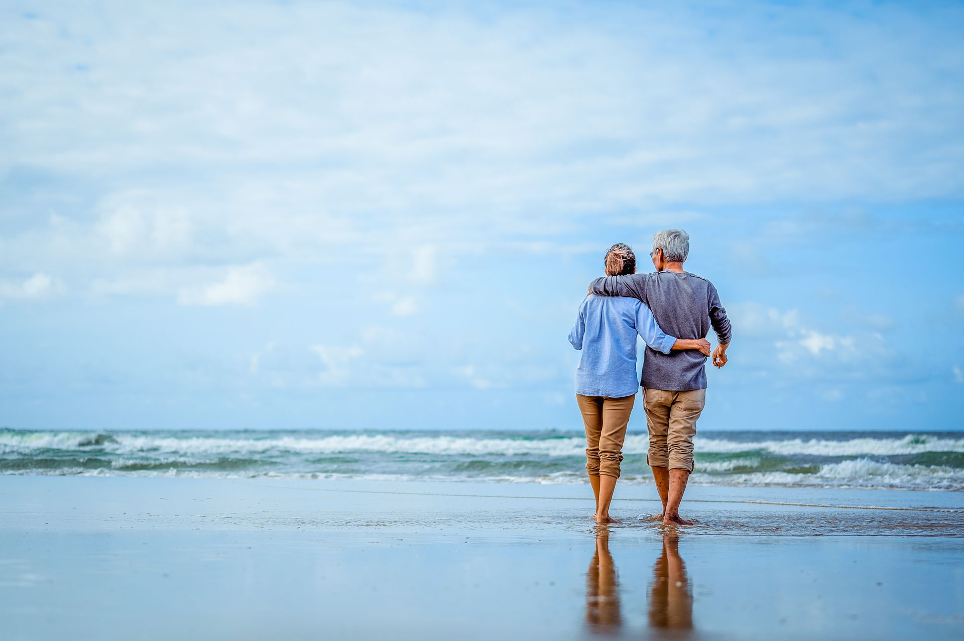 Two people with arms around each other walking on a sandy beach toward the ocean under a blue, cloudy sky.
