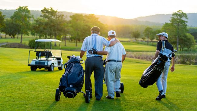 Four golfers walk across a sunny green course, carrying bags and pulling a cart at sunrise.