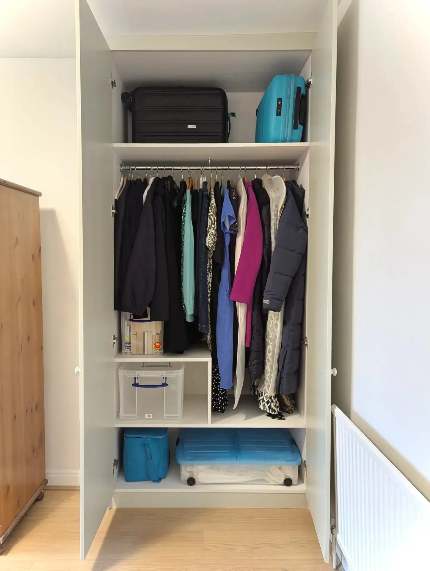 White wardrobe open, displaying hanging clothes and storage boxes, with suitcases overhead.