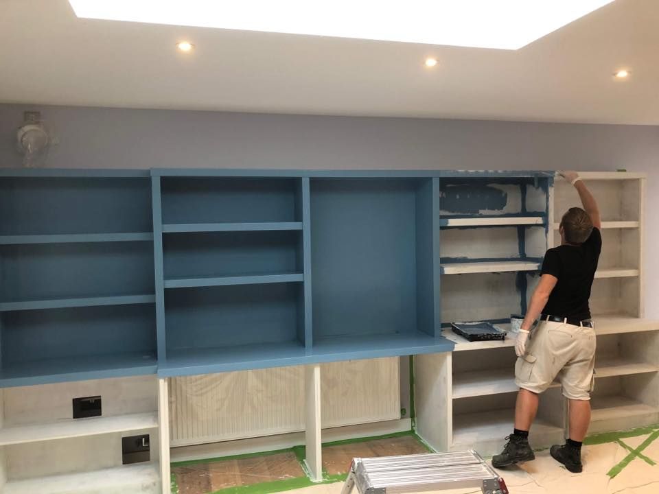 Person painting a built-in bookshelf blue. The room has a skylight and light blue walls.