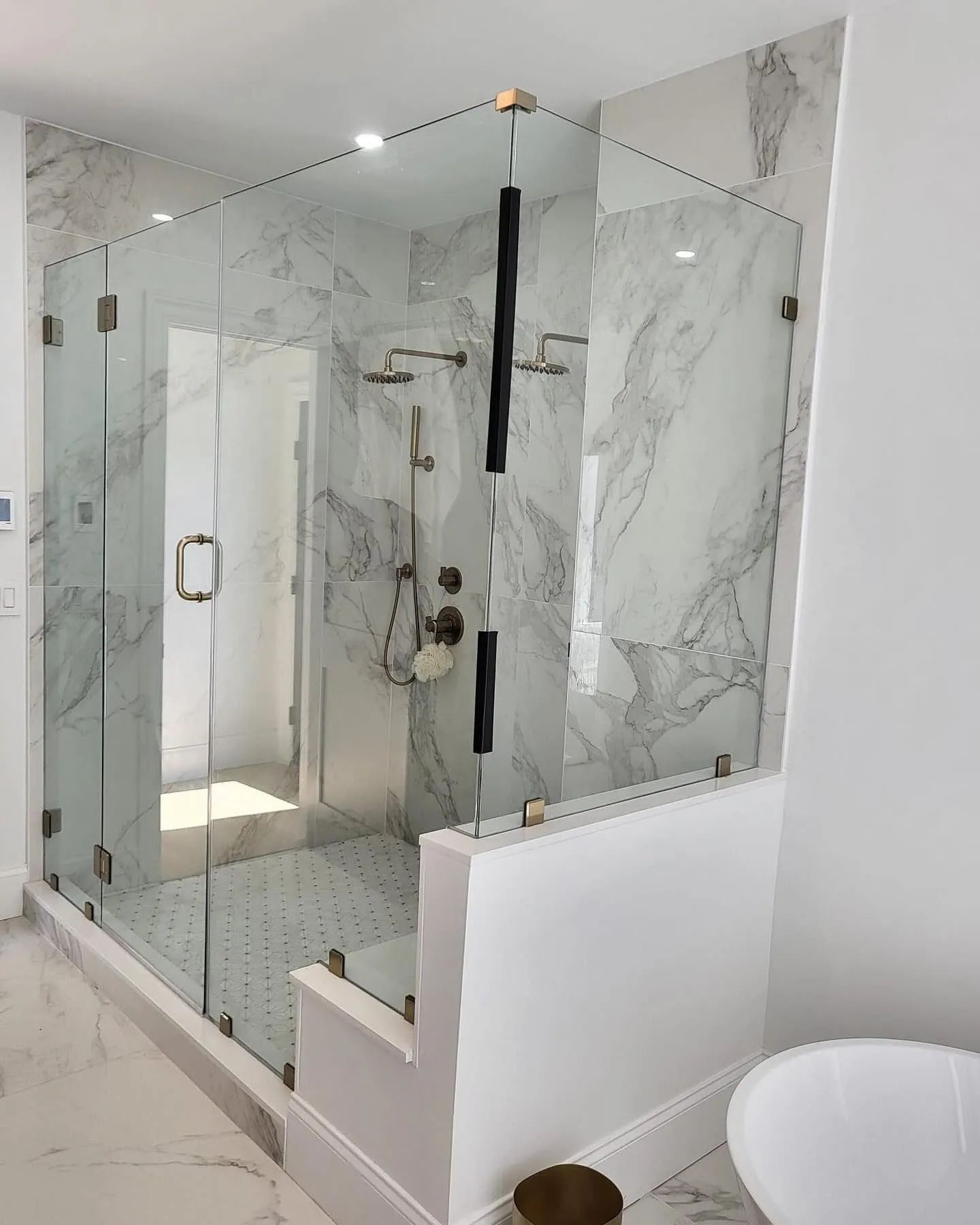 Glass-enclosed marble shower with gold fixtures in a bright bathroom, next to a white bathtub