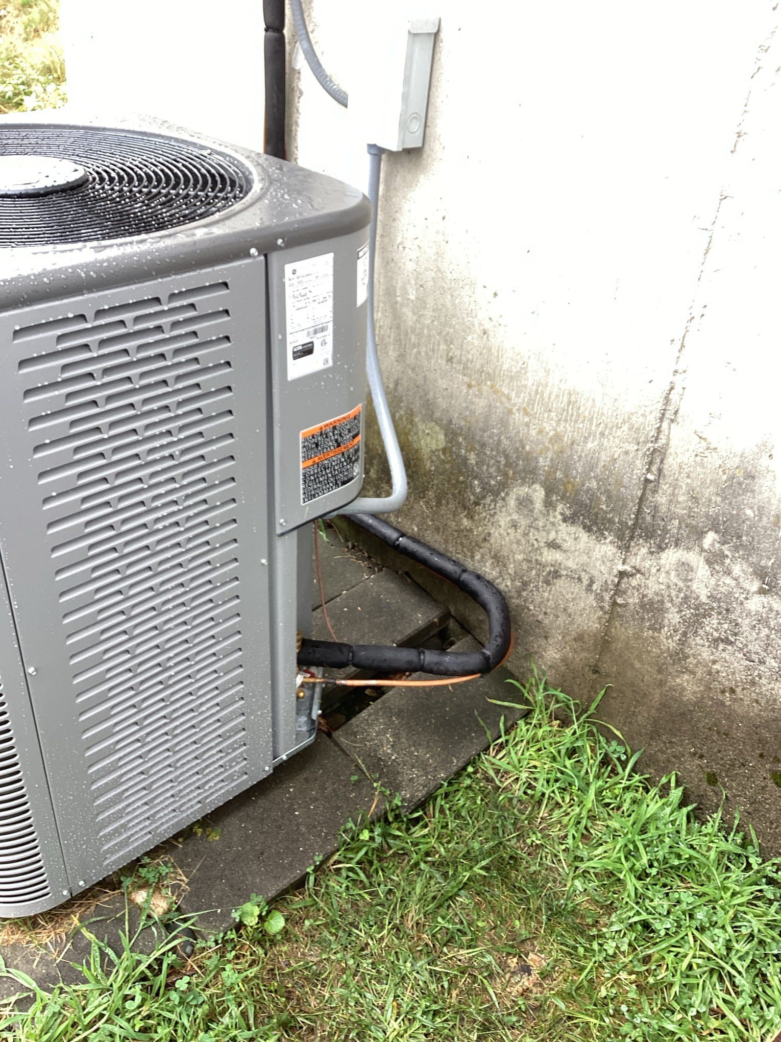 Gray air conditioning unit next to a concrete wall and patch of grass. Copper tubes visible.