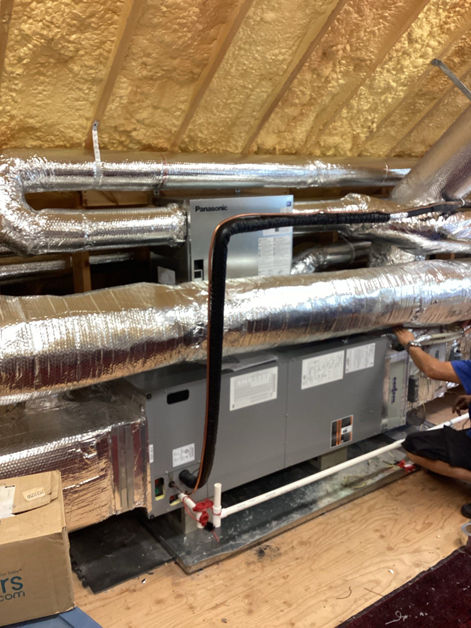 HVAC system installation in an attic. A person works on the unit, surrounded by ductwork and insulation.