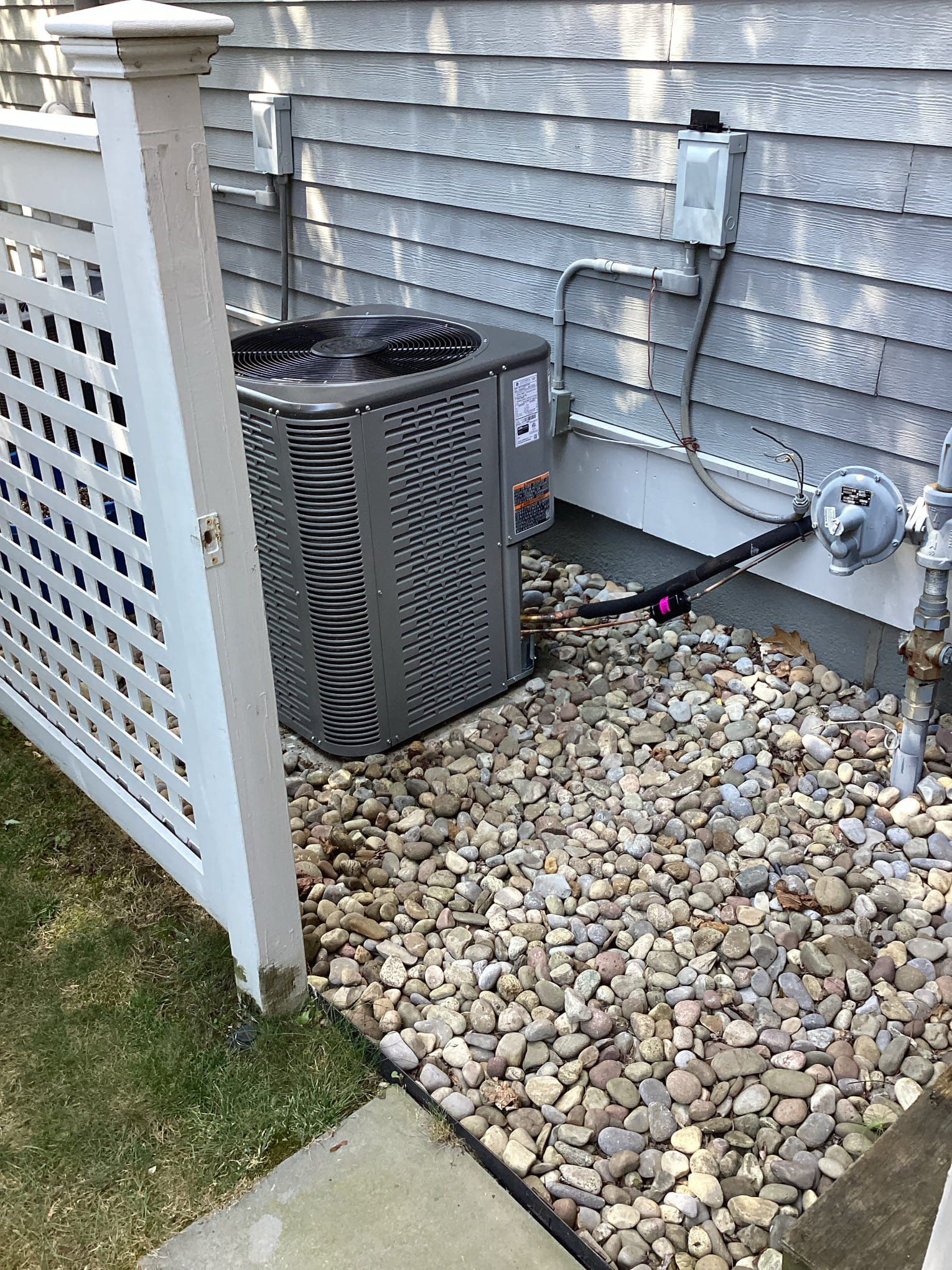 Air conditioning unit next to a white fence and a gray house with rocks.