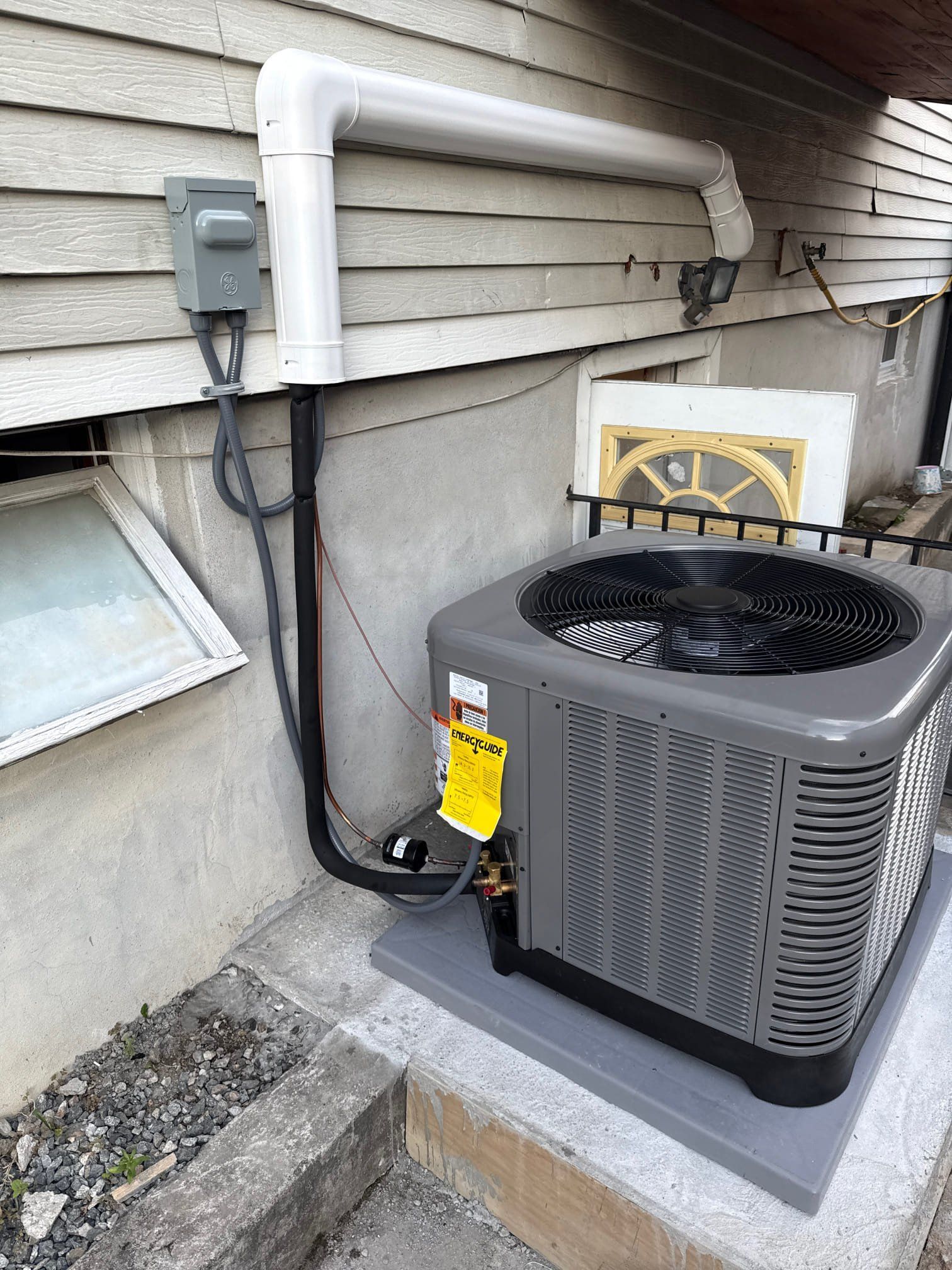Exterior air conditioning unit next to a building with electrical conduit and a window.