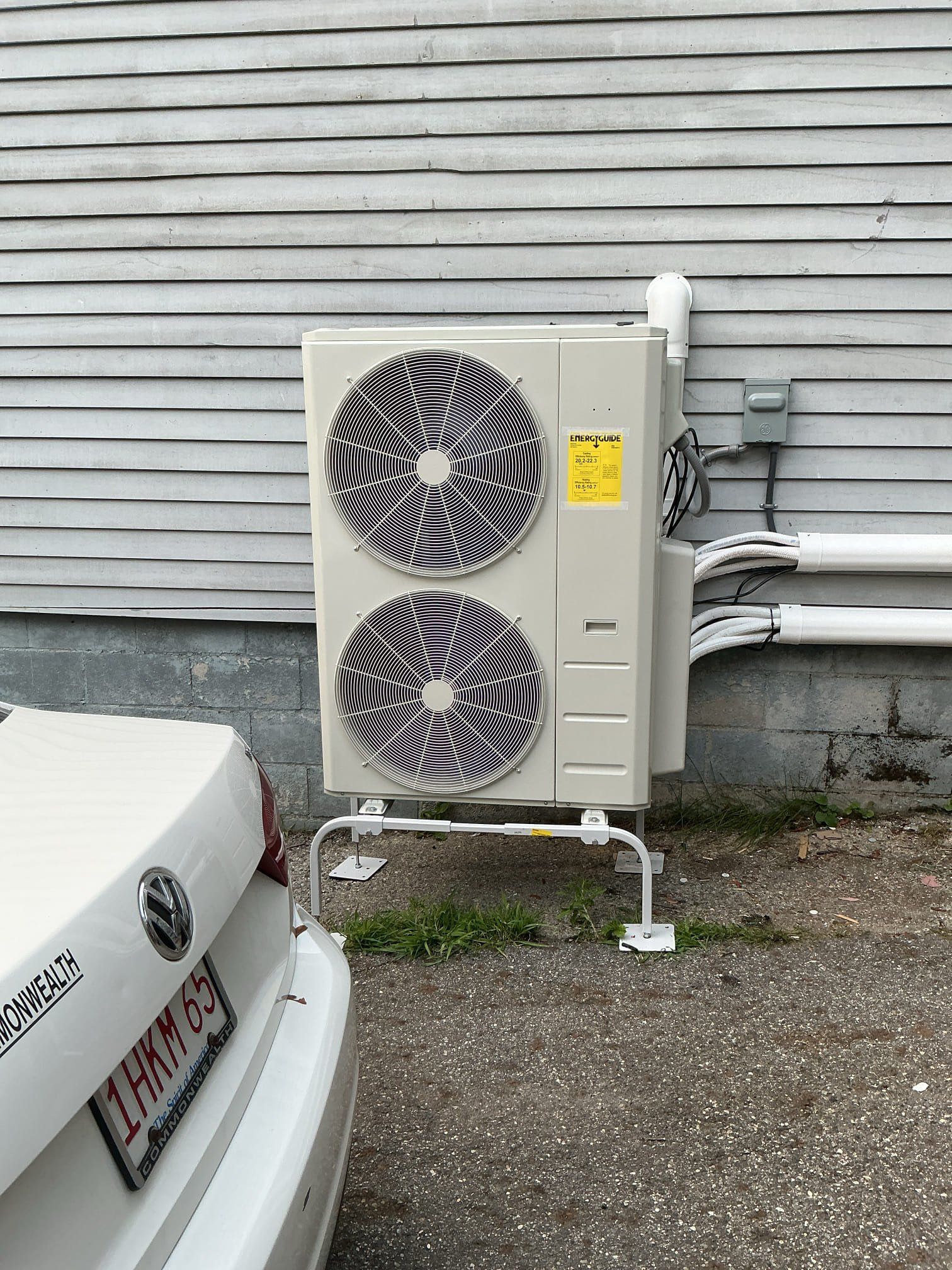 White heat pump unit on a metal stand outside a building with white siding, near a white car.