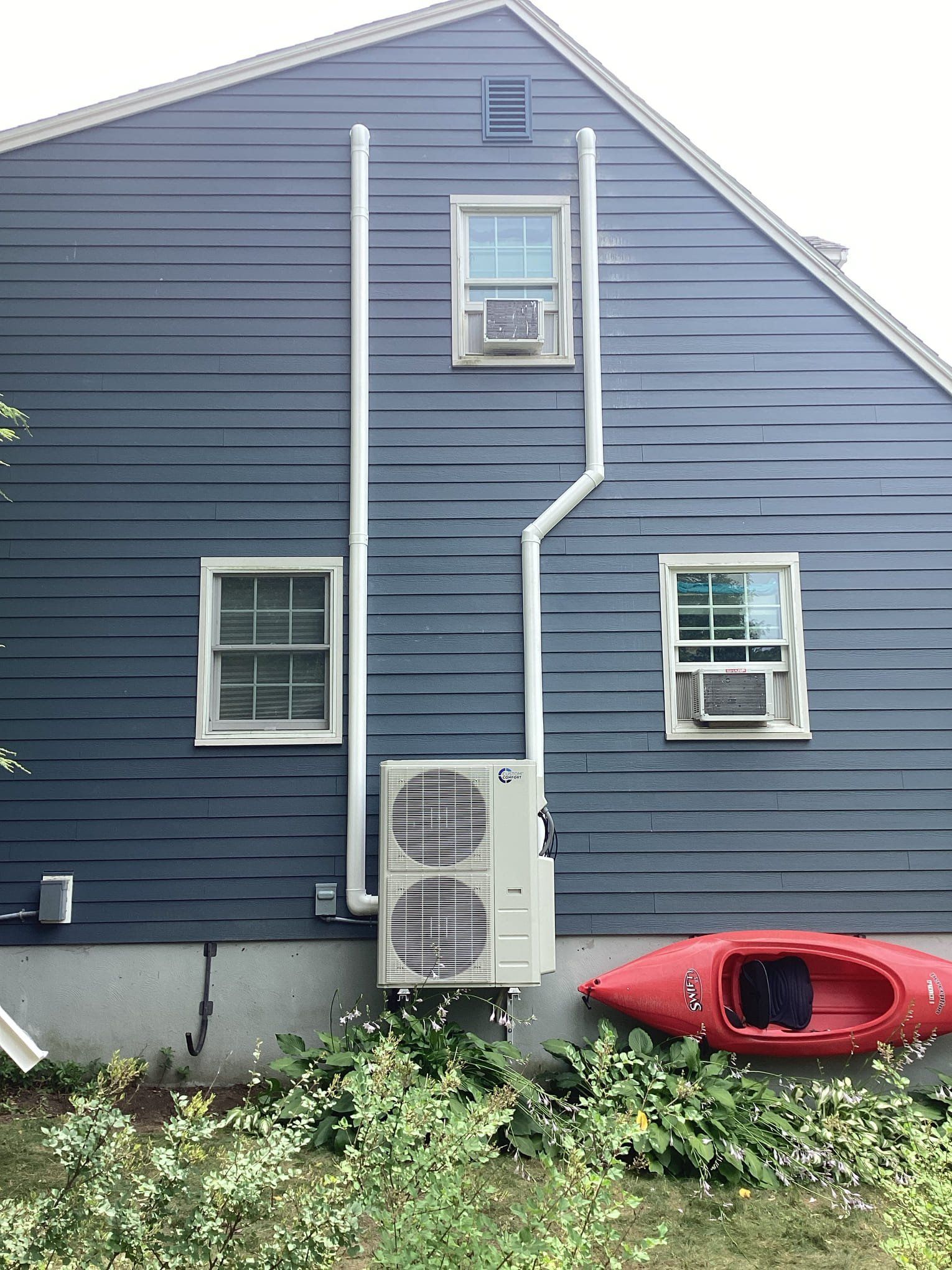 Exterior of a blue house with two air conditioners. White pipes run up the side of the house to the windows. A red kayak rests below.