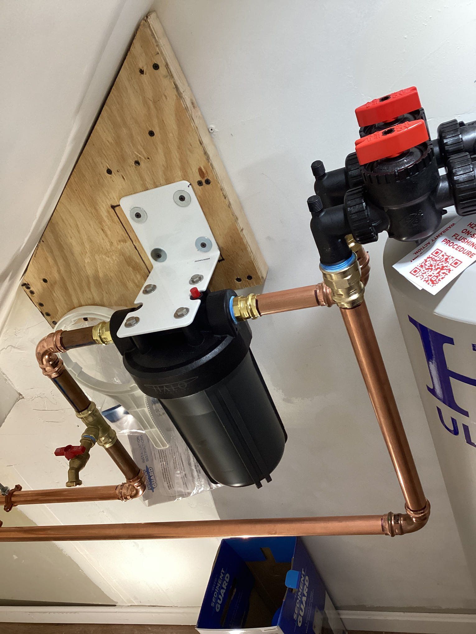 Water filtration system with black filter housing, copper pipes, and red valve caps mounted on a wooden board.