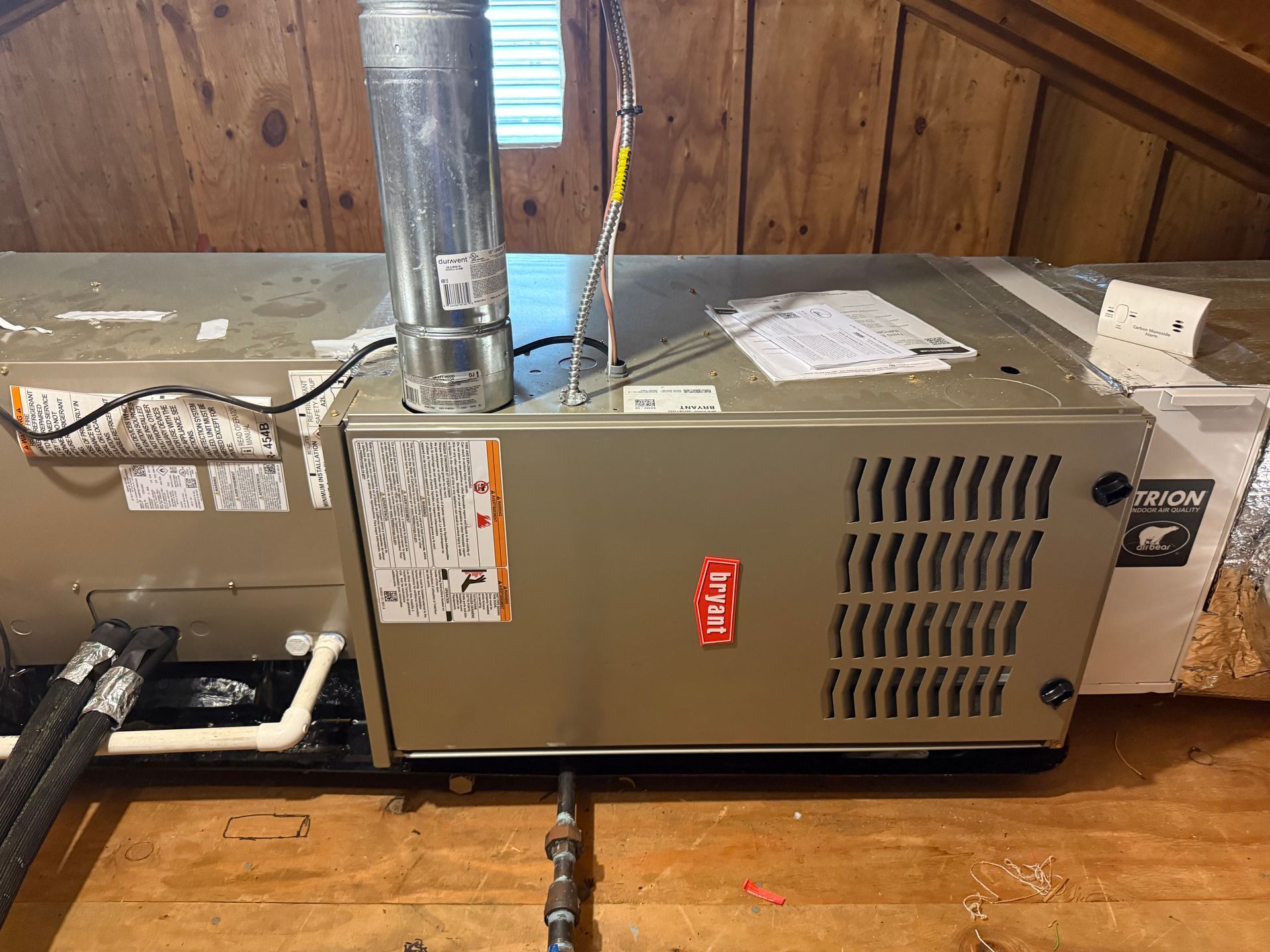 Tan furnace in an attic with ductwork and a metal vent pipe.