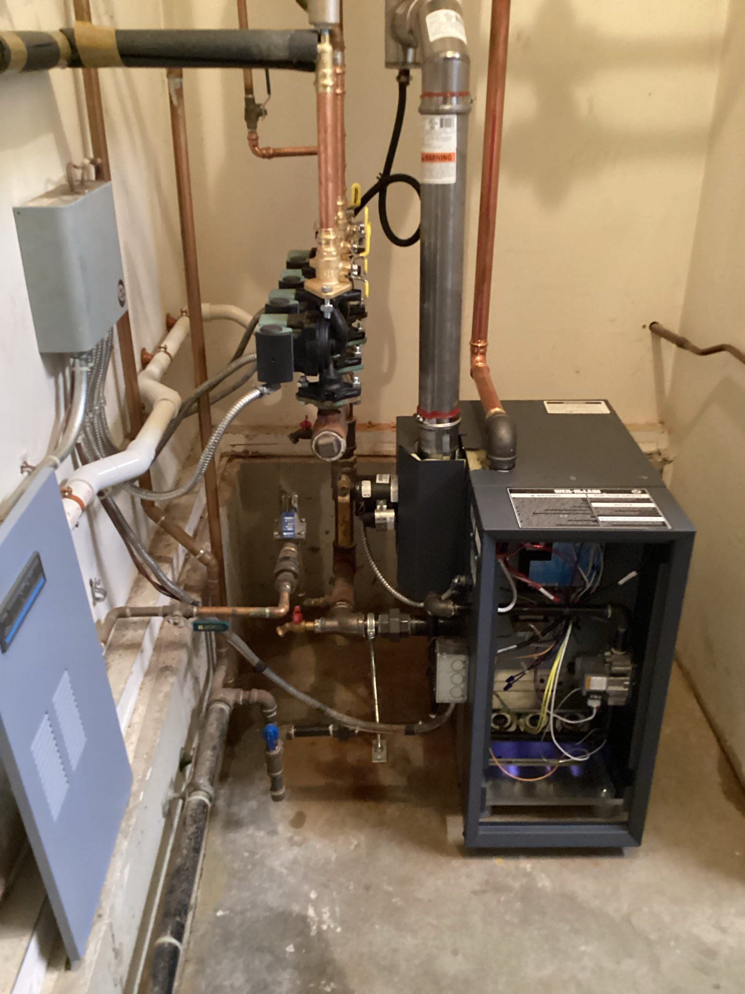 A residential heating system with copper pipes, valves, and a gray metal cabinet, installed against a wall.