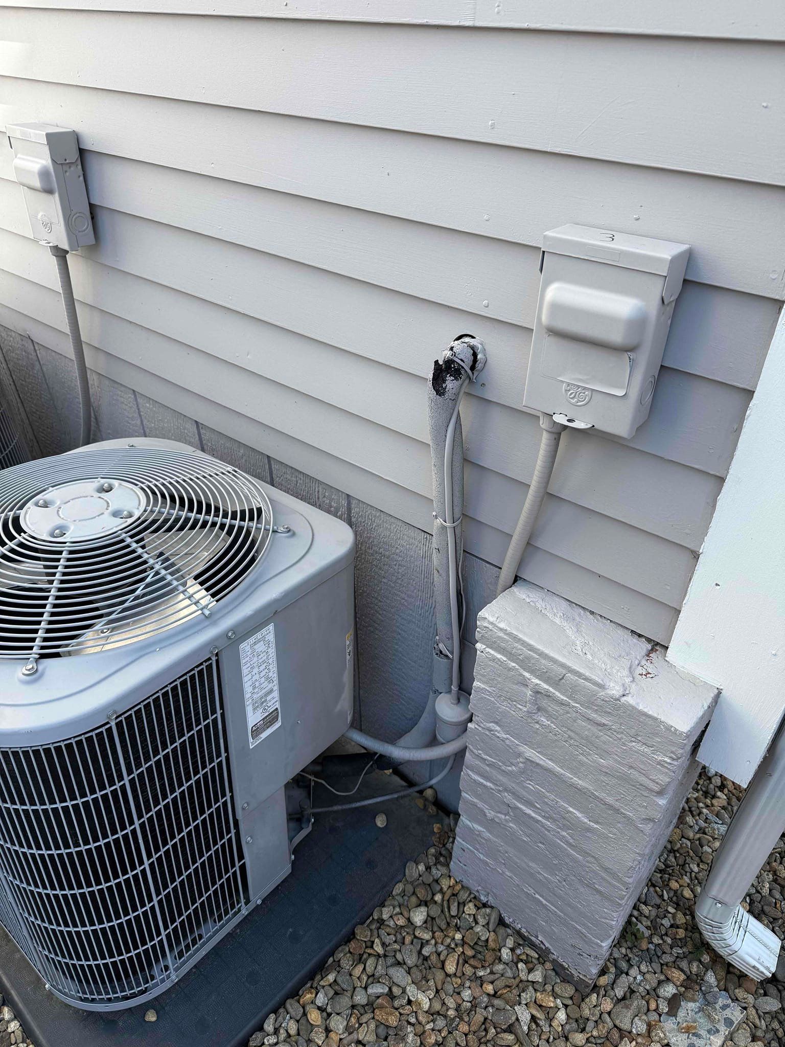 Air conditioning unit outside a light gray building; electrical components visible.
