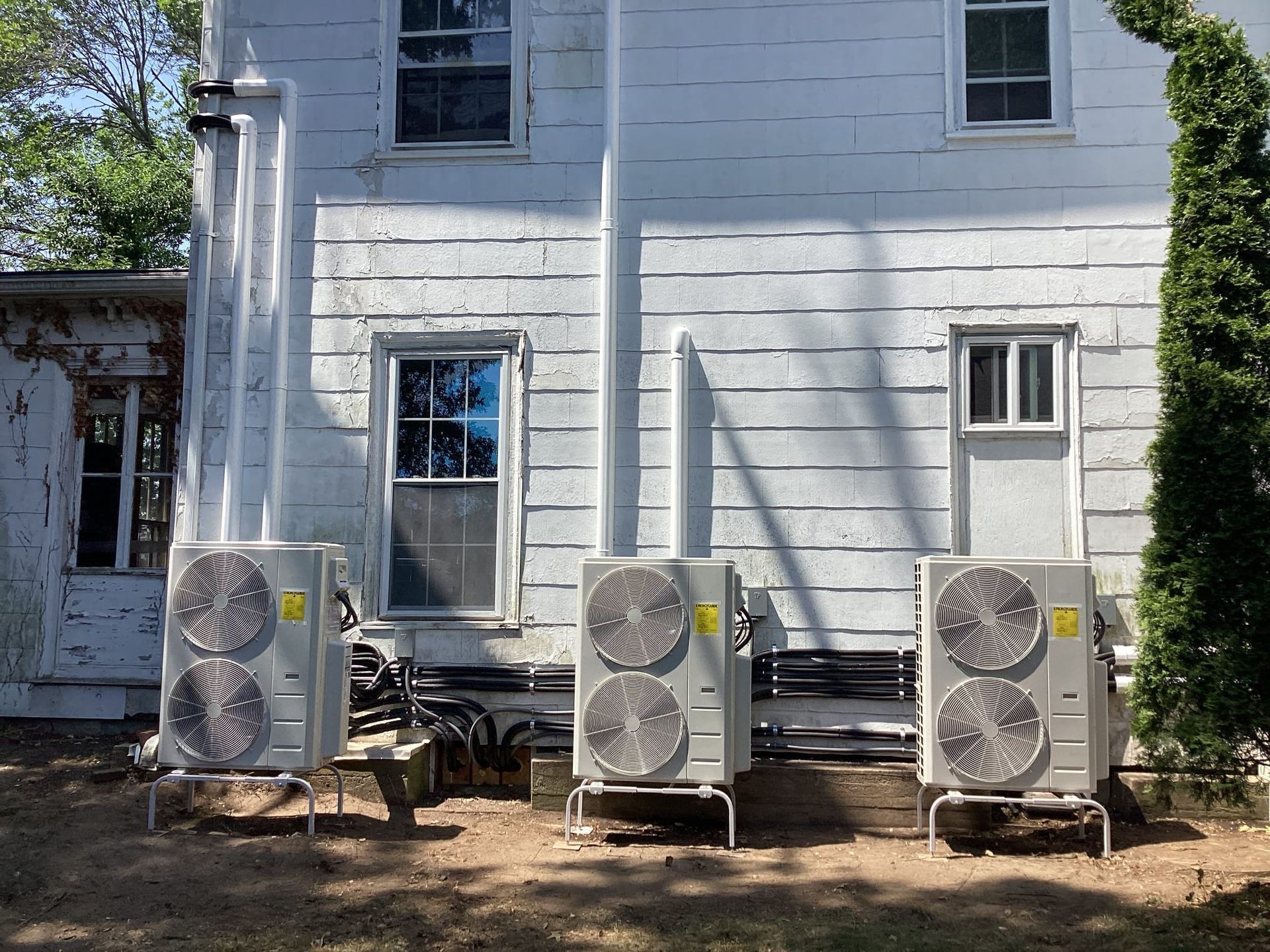 Three outdoor air conditioning units on stands next to a white house with windows.