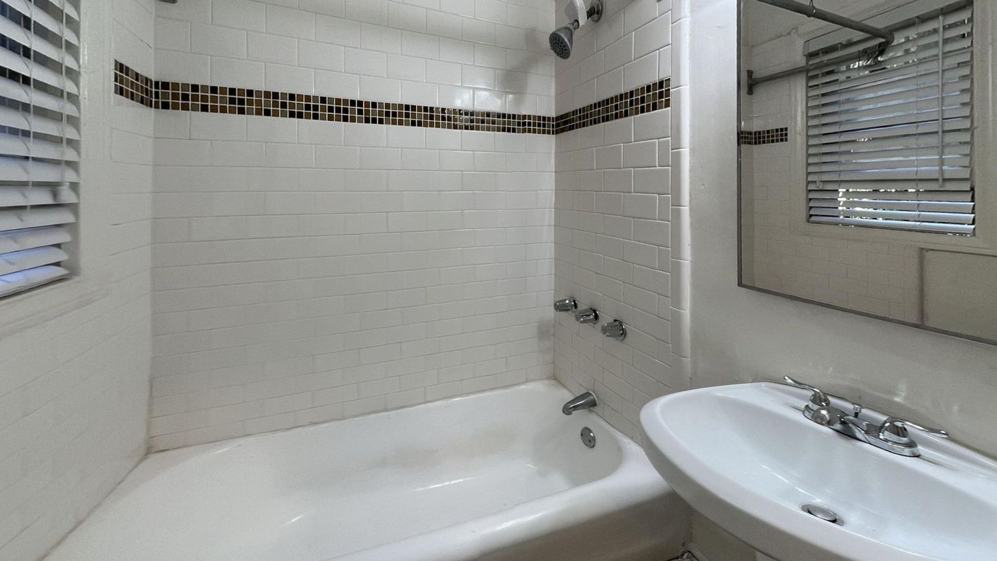 Bathroom with white tiled walls, bathtub, sink, and window with blinds.