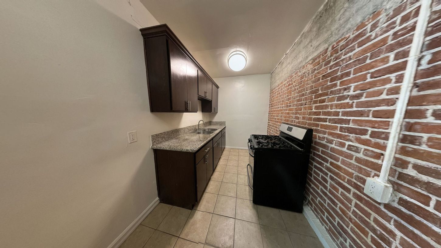 Narrow kitchen with dark cabinets, brick wall, and stove.