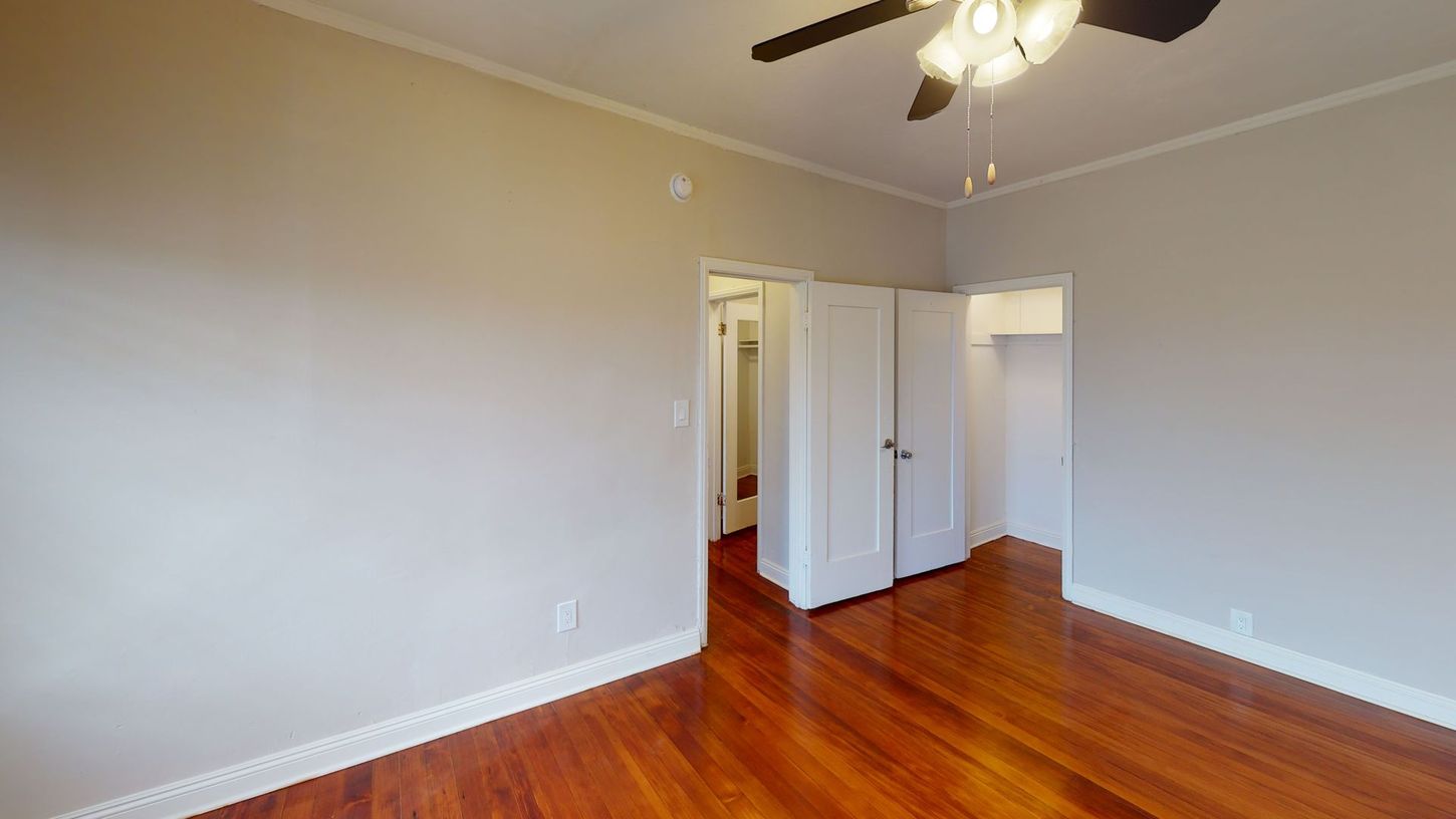 Empty bedroom with hardwood floors, two doors, and a ceiling fan. Walls are painted light gray.