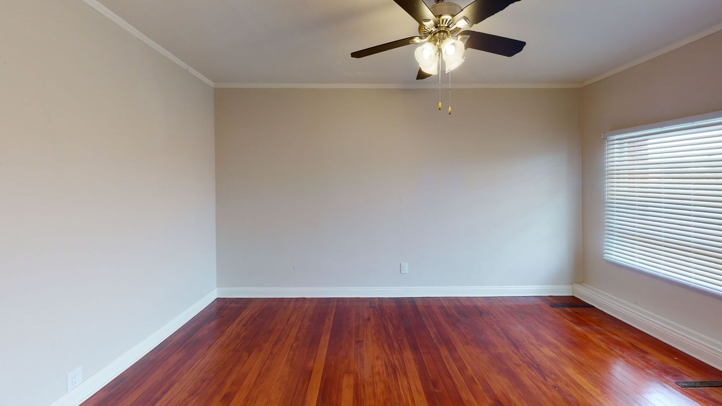Empty room with hardwood floors, beige walls, a window with blinds, and a ceiling fan.