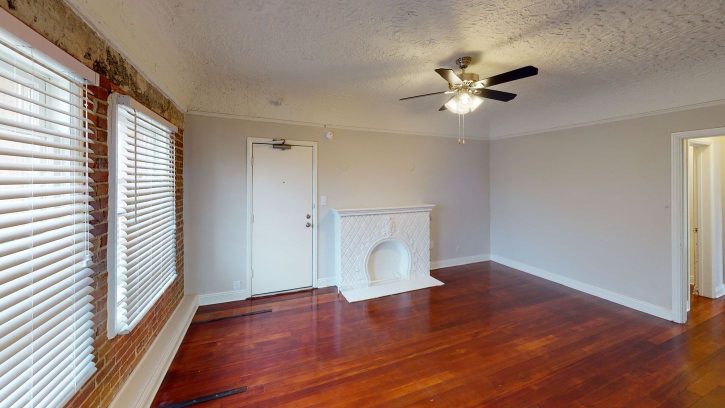 Living room with dark wood floors, white fireplace, windows with blinds, brick wall.