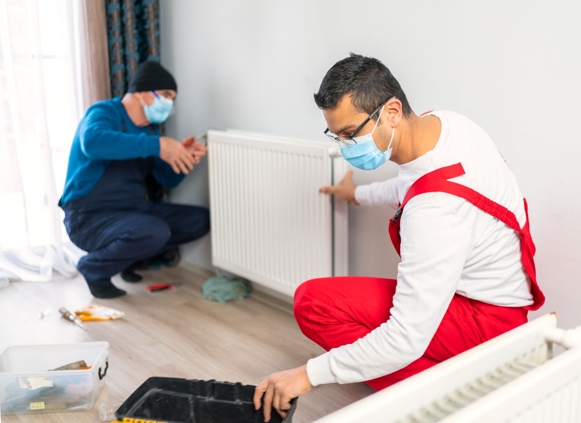 Two professional plumbers with protective face mask are fixing heating radiator in living room | Wel