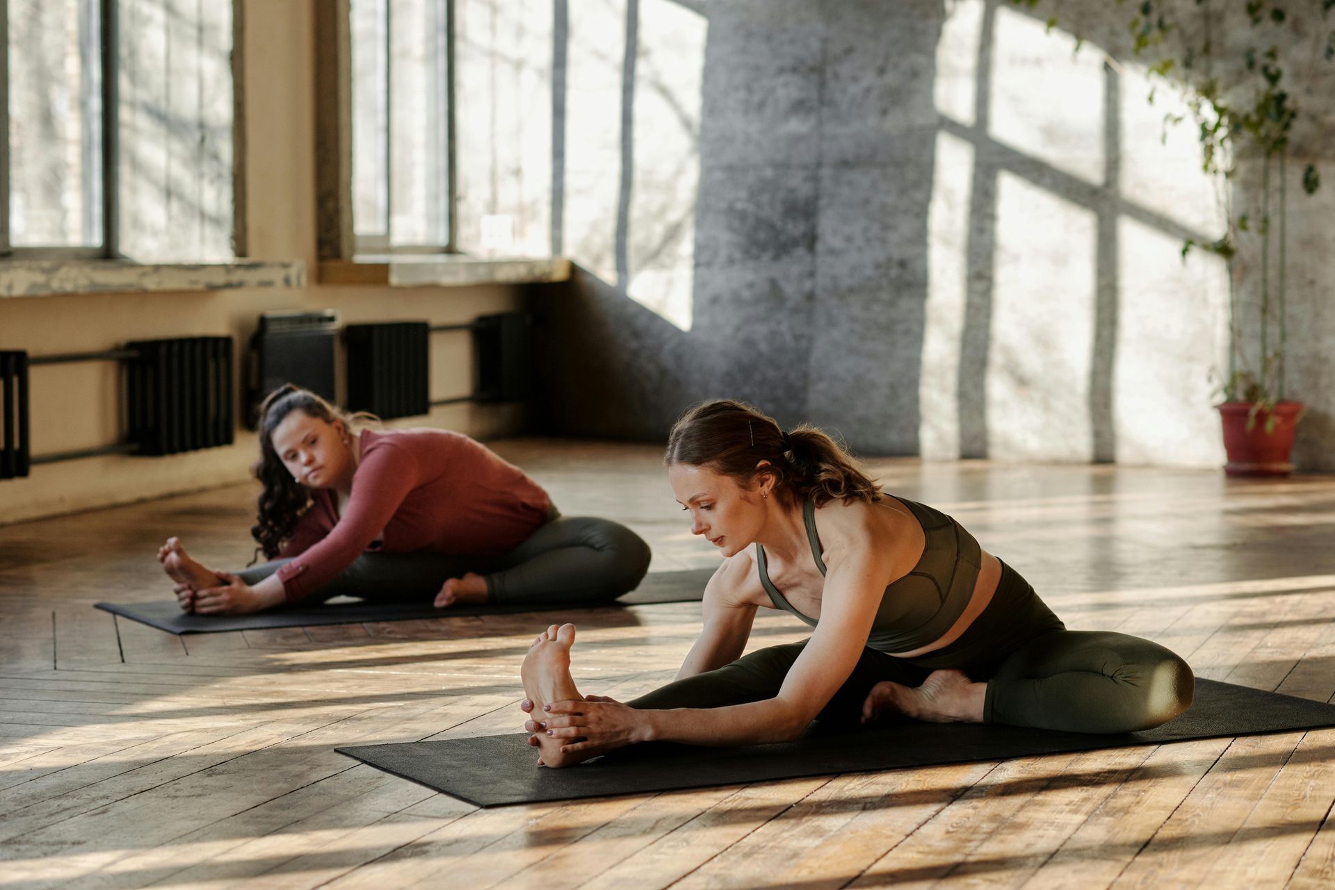 Two people stretching on yoga mats in a sunlit studio, reaching towards their feet.
