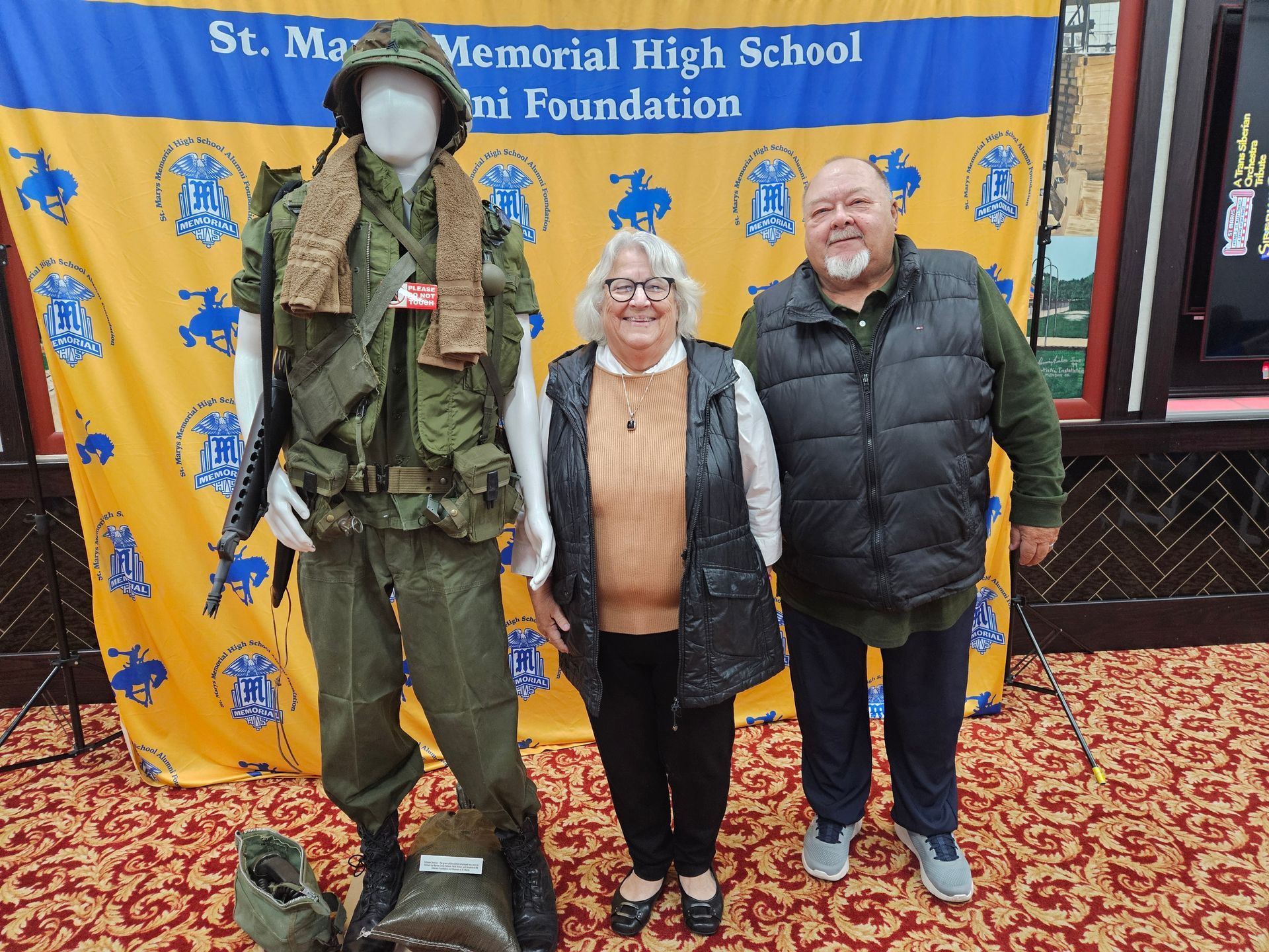 Two people pose next to a military uniform display with a school banner.