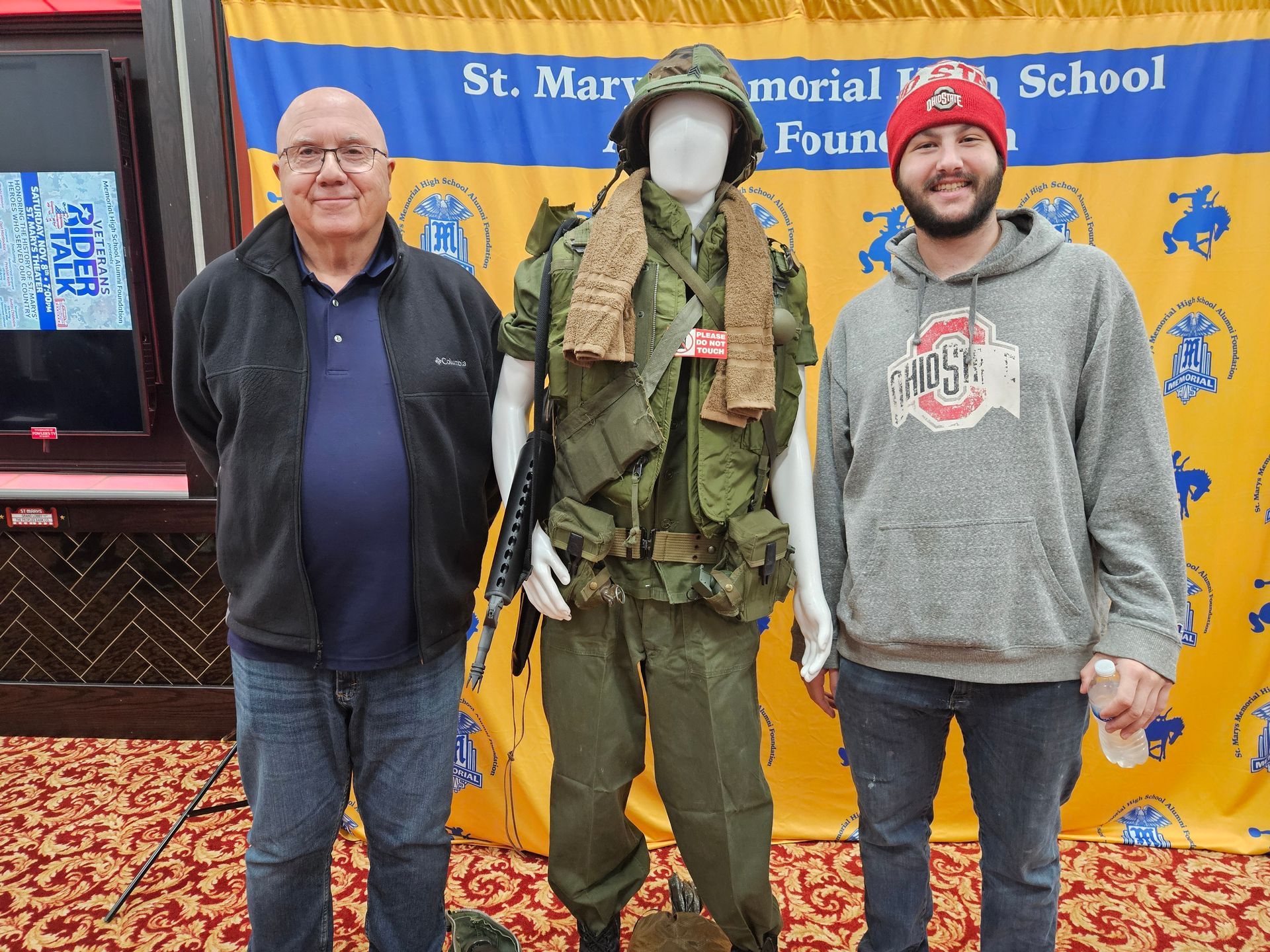 Two men stand by a mannequin dressed in military fatigues; blue and gold banner in the background.