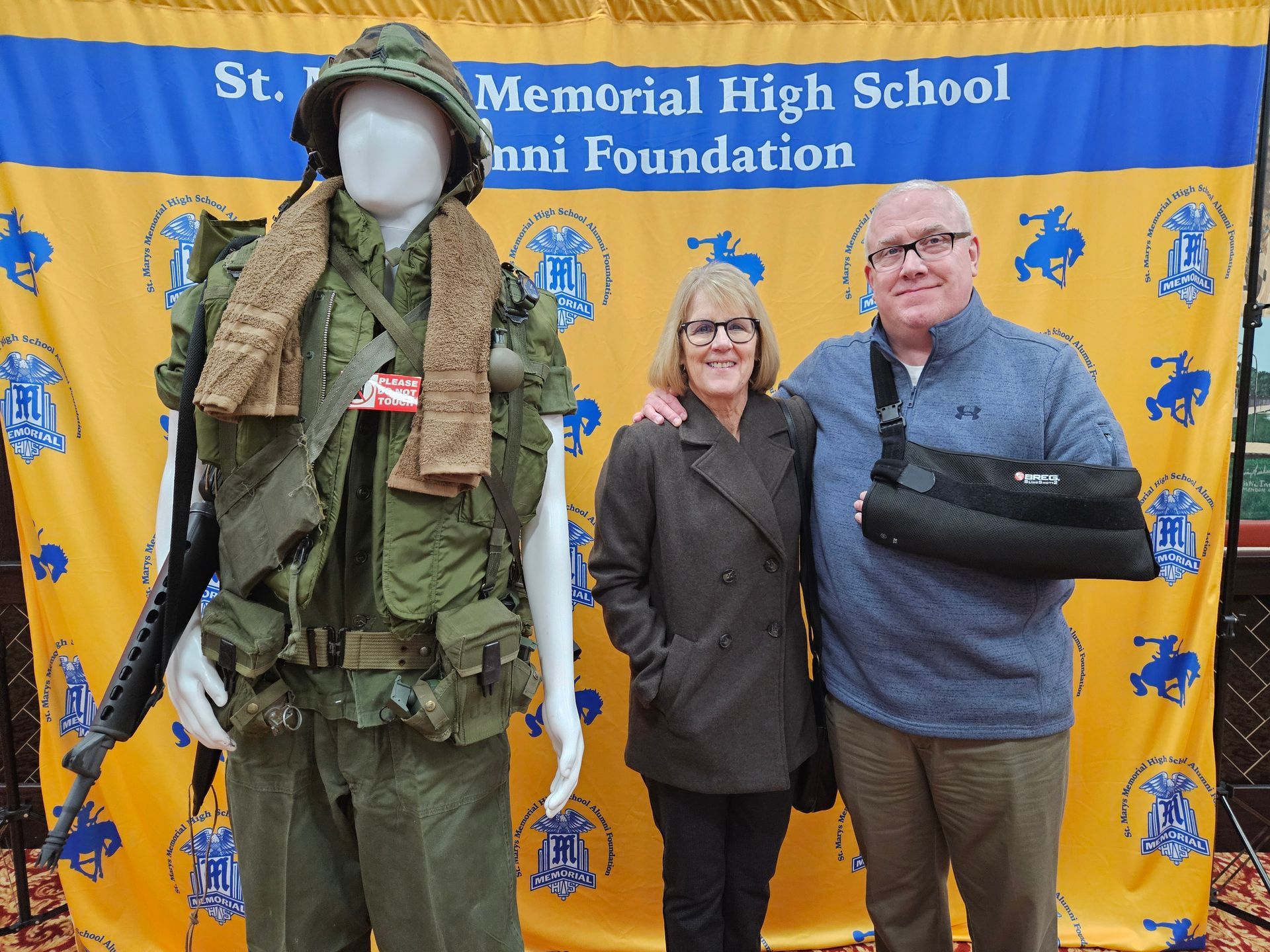 A man with a sling and a woman pose next to a mannequin in a military uniform. Behind them is a school banner.