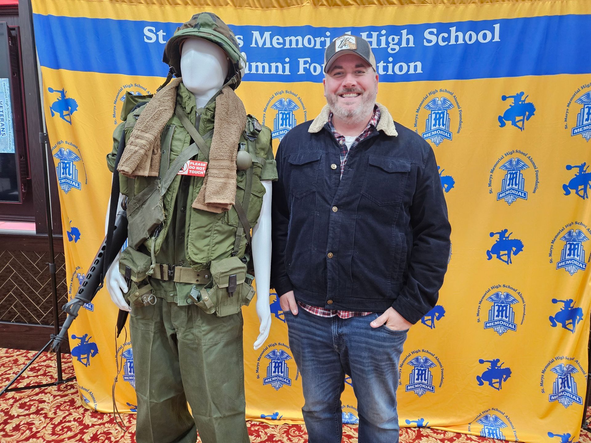 Man stands by a mannequin in Vietnam War uniform, in front of a banner for an alumni foundation.