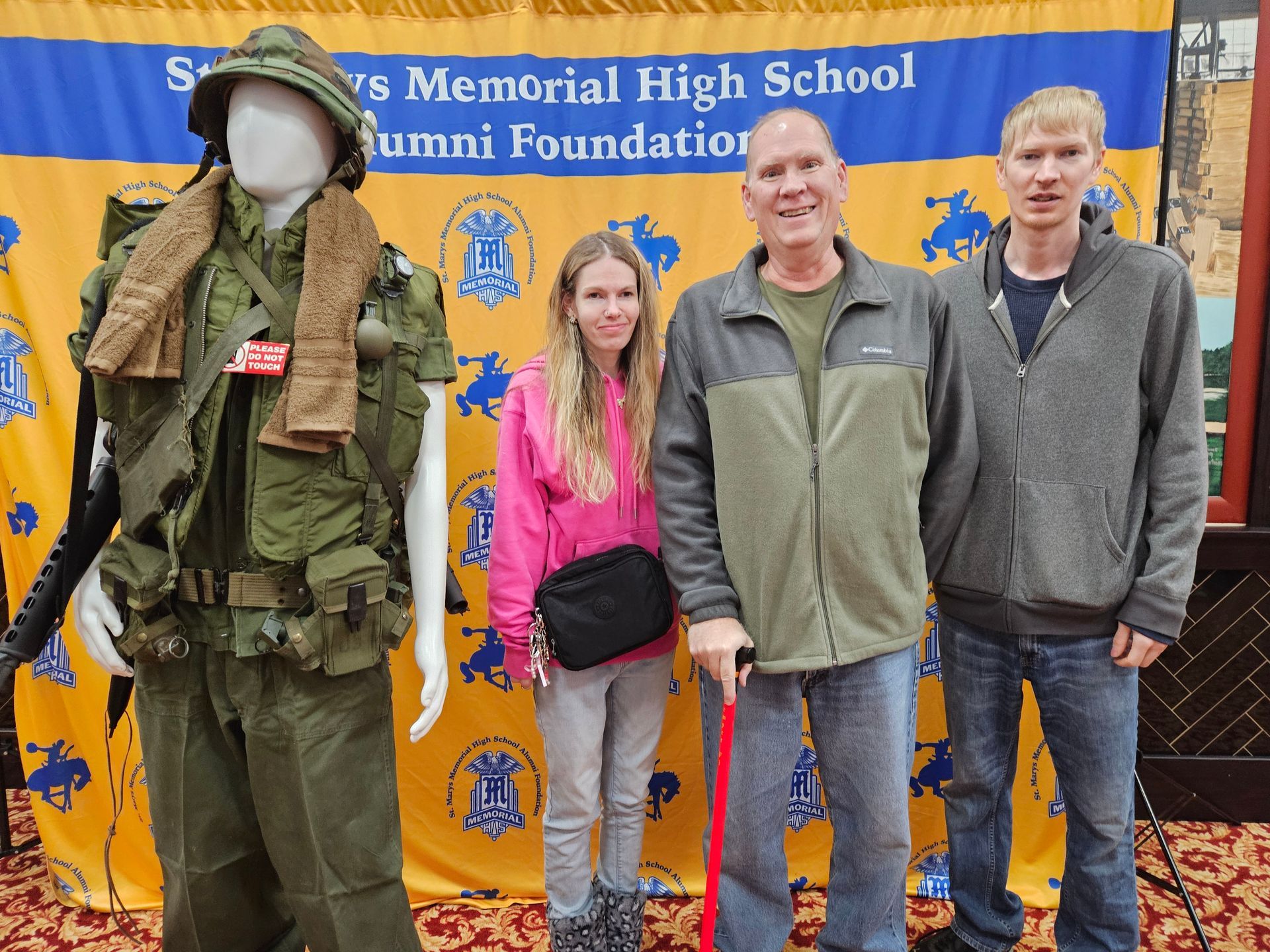 People pose by a Vietnam War uniform display at a memorial high school alumni foundation event.