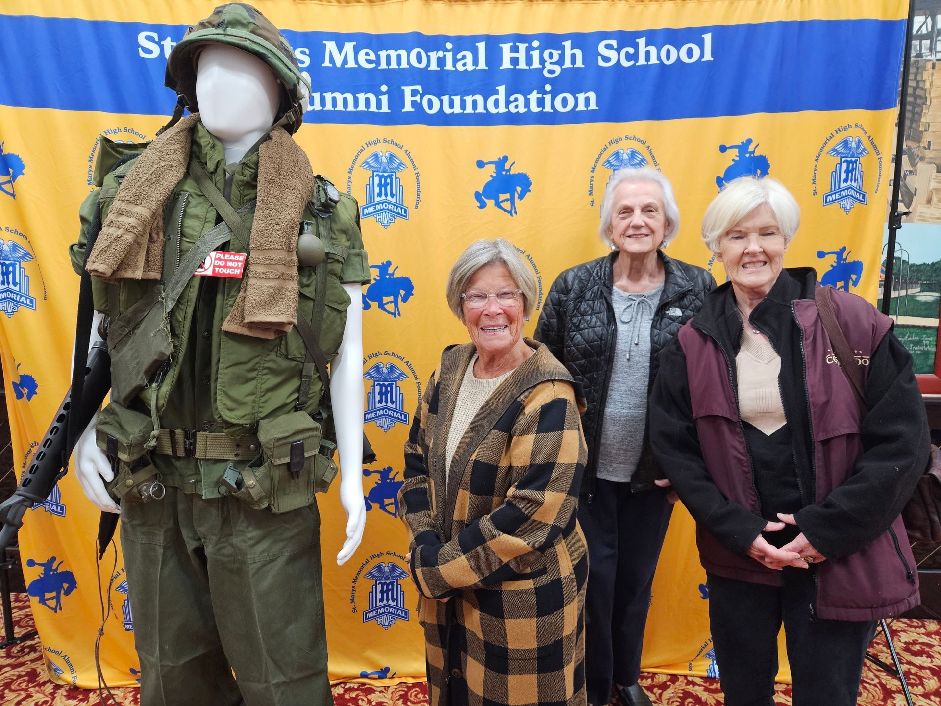 Three women pose in front of a backdrop with a military uniform display.