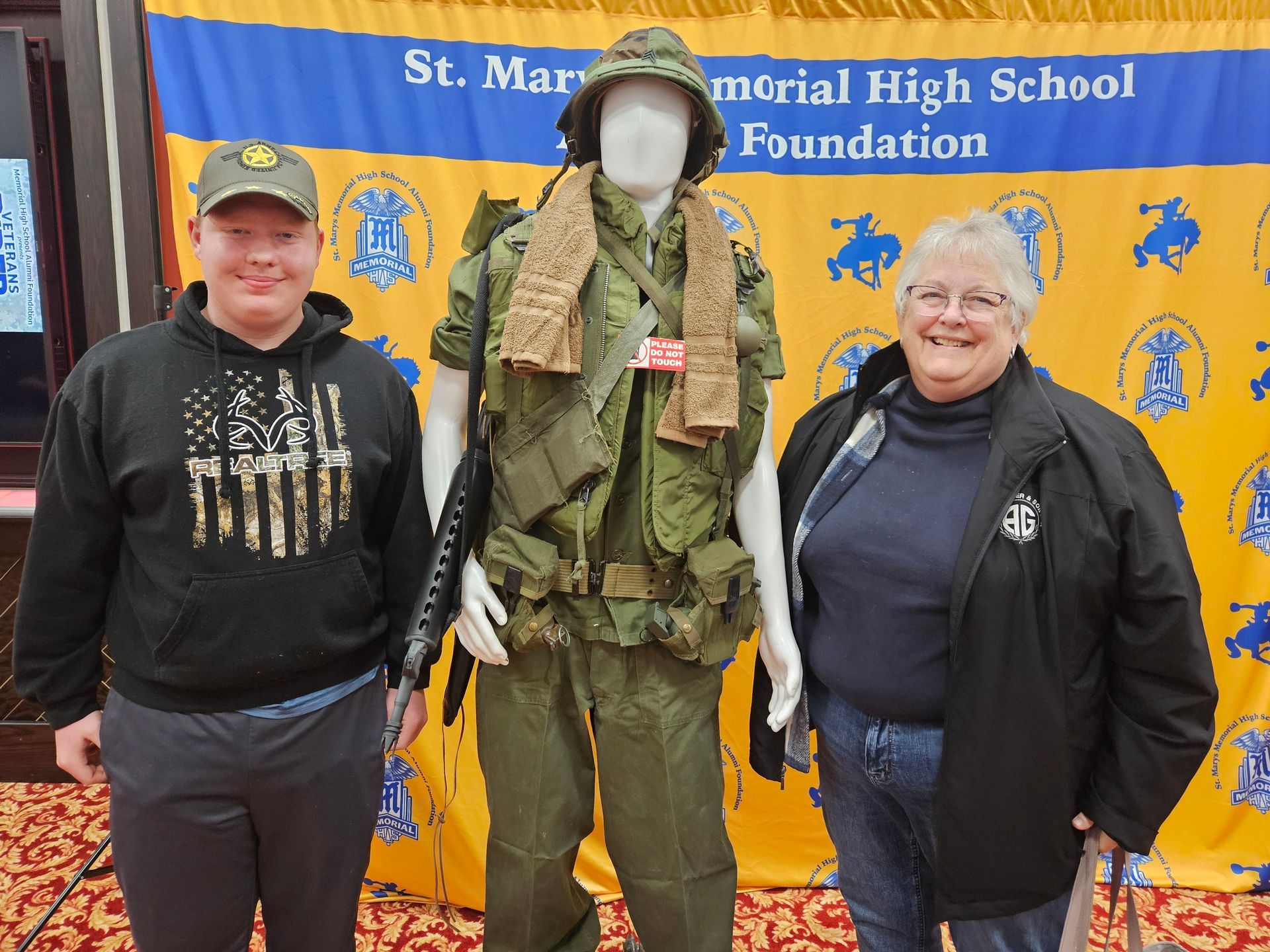 Two people pose with a mannequin in military uniform. St. Marys Memorial High School Foundation banner in background.