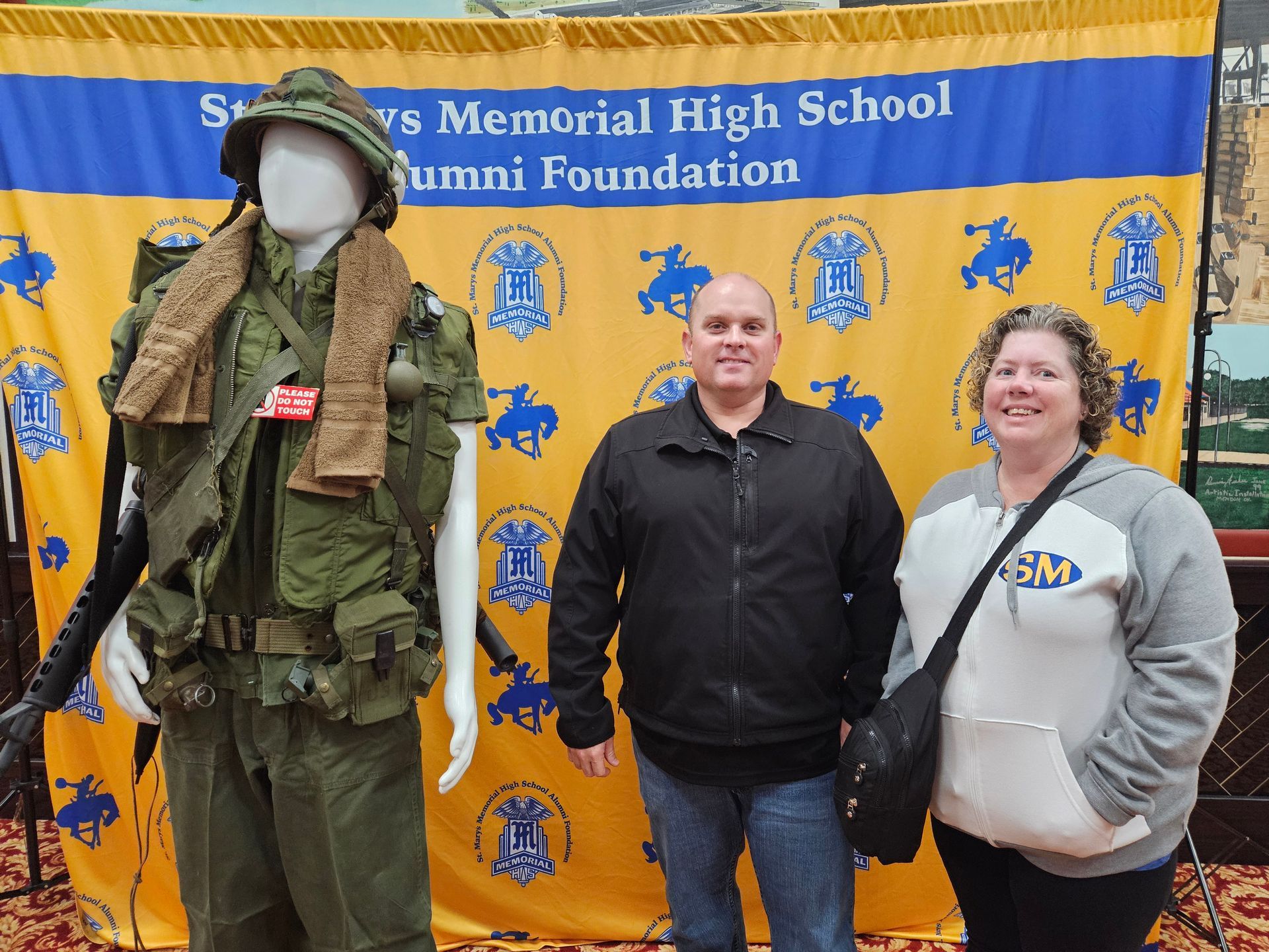 Man and woman stand next to a mannequin in a military uniform in front of a school alumni backdrop.