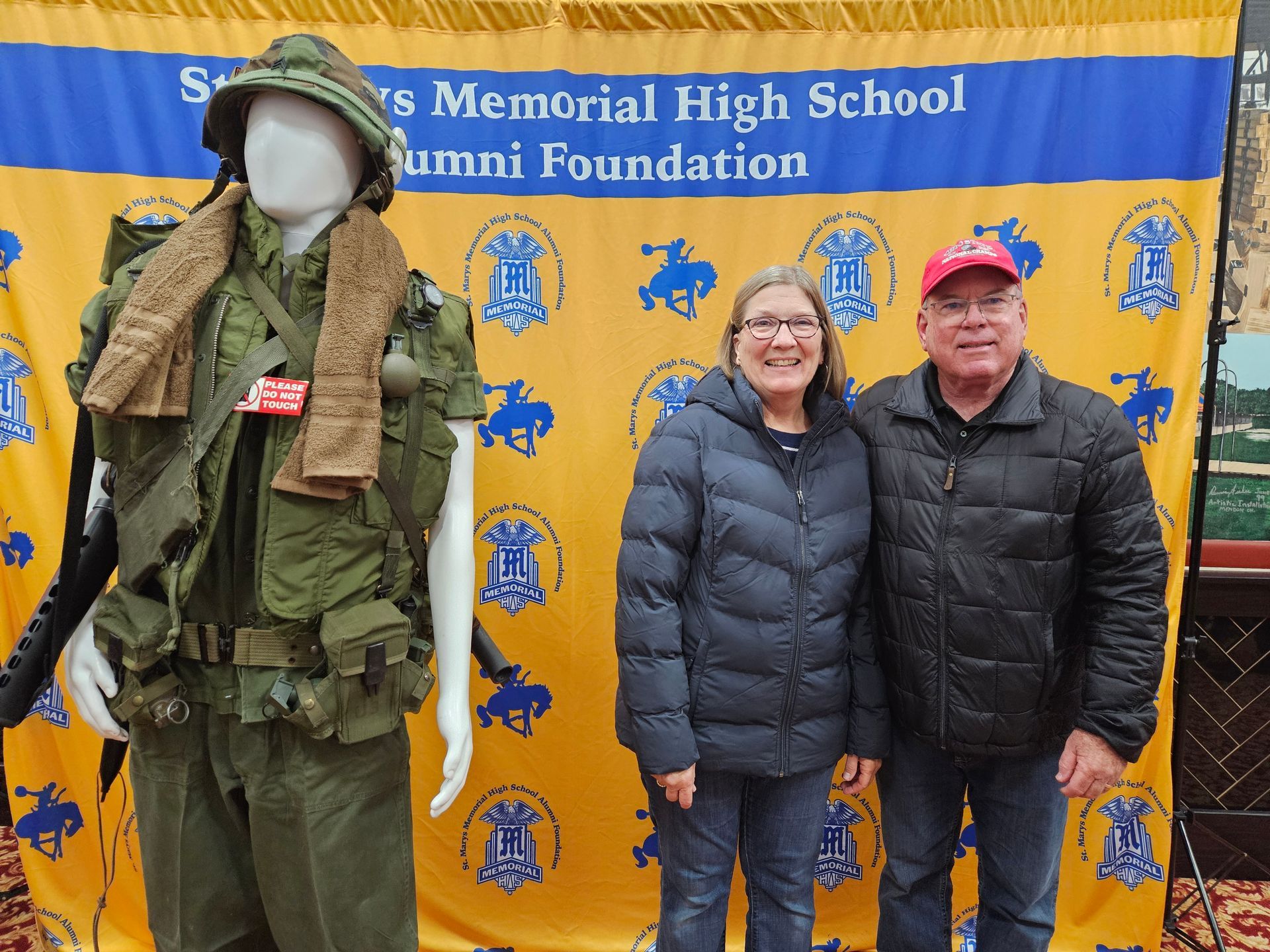 A couple stands near a mannequin in military fatigues and a banner for an alumni foundation.
