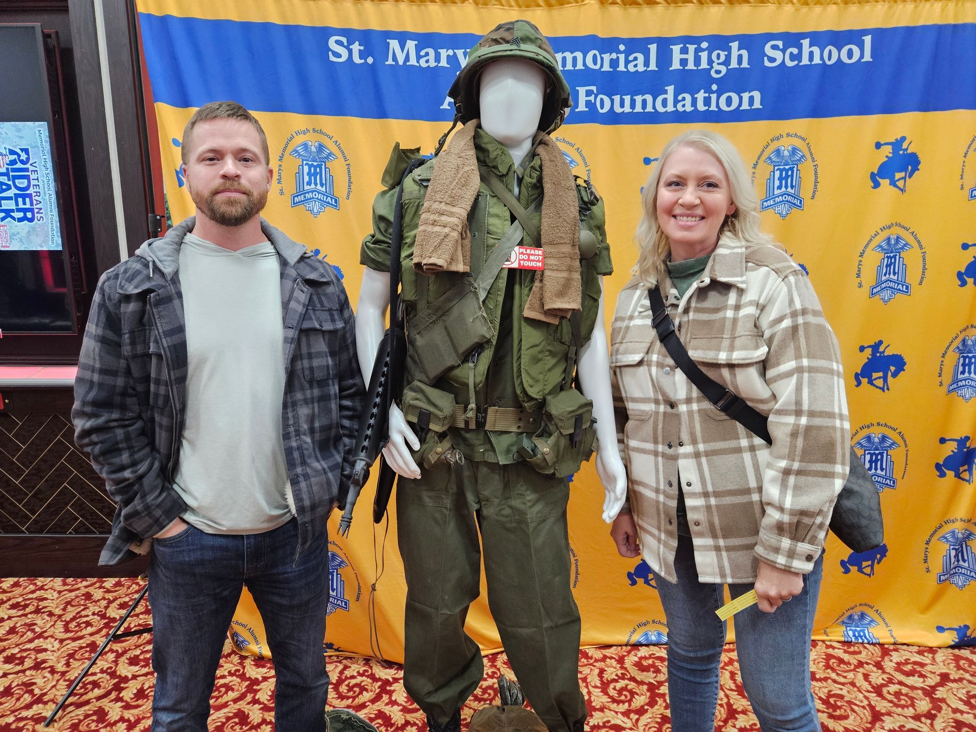 Two people stand beside a military uniform on display. Background: St. Mary's Memorial High School Foundation banner.