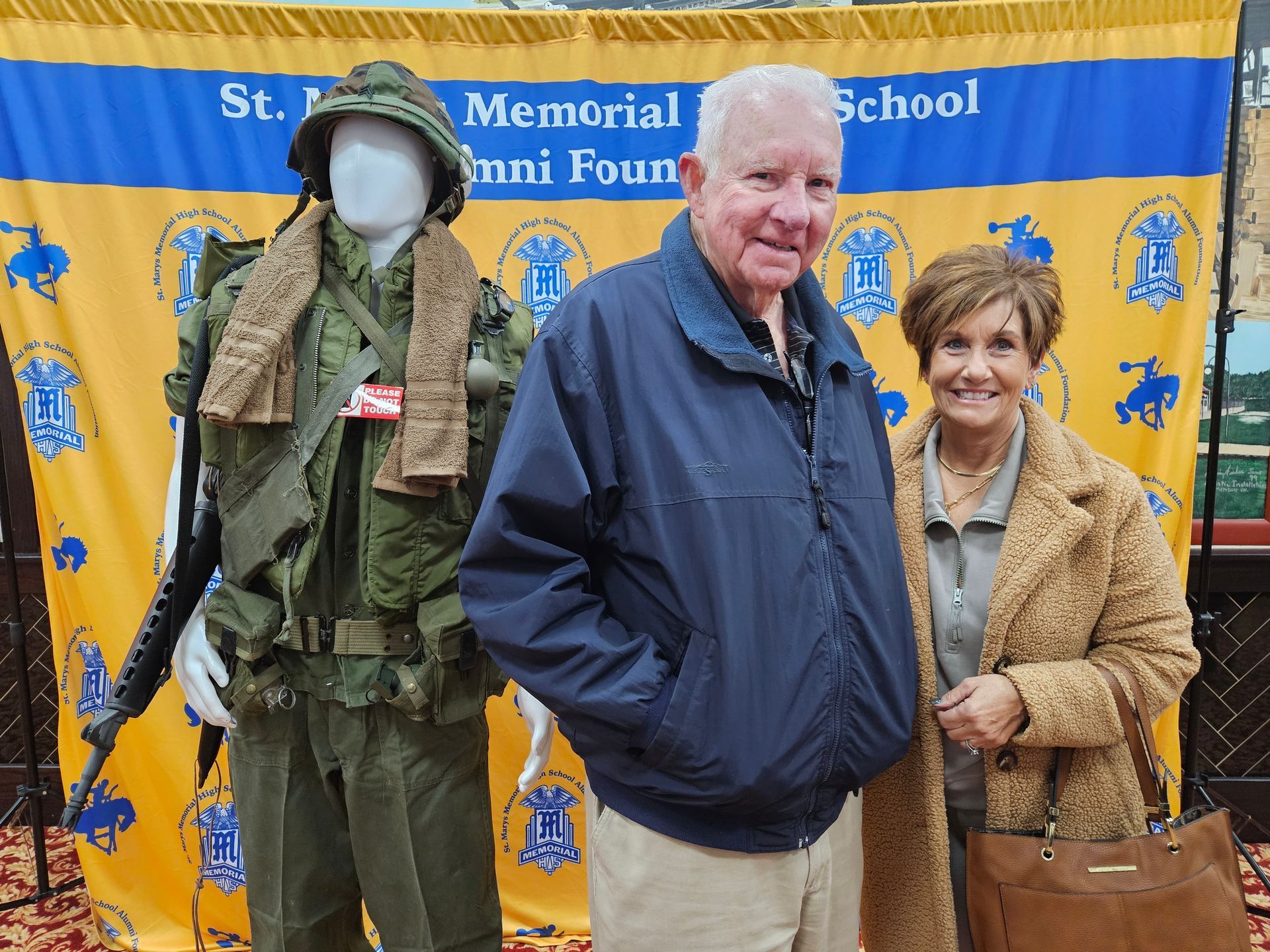 Man and woman stand by a mannequin in military uniform. They smile, in front of a school alumni banner.