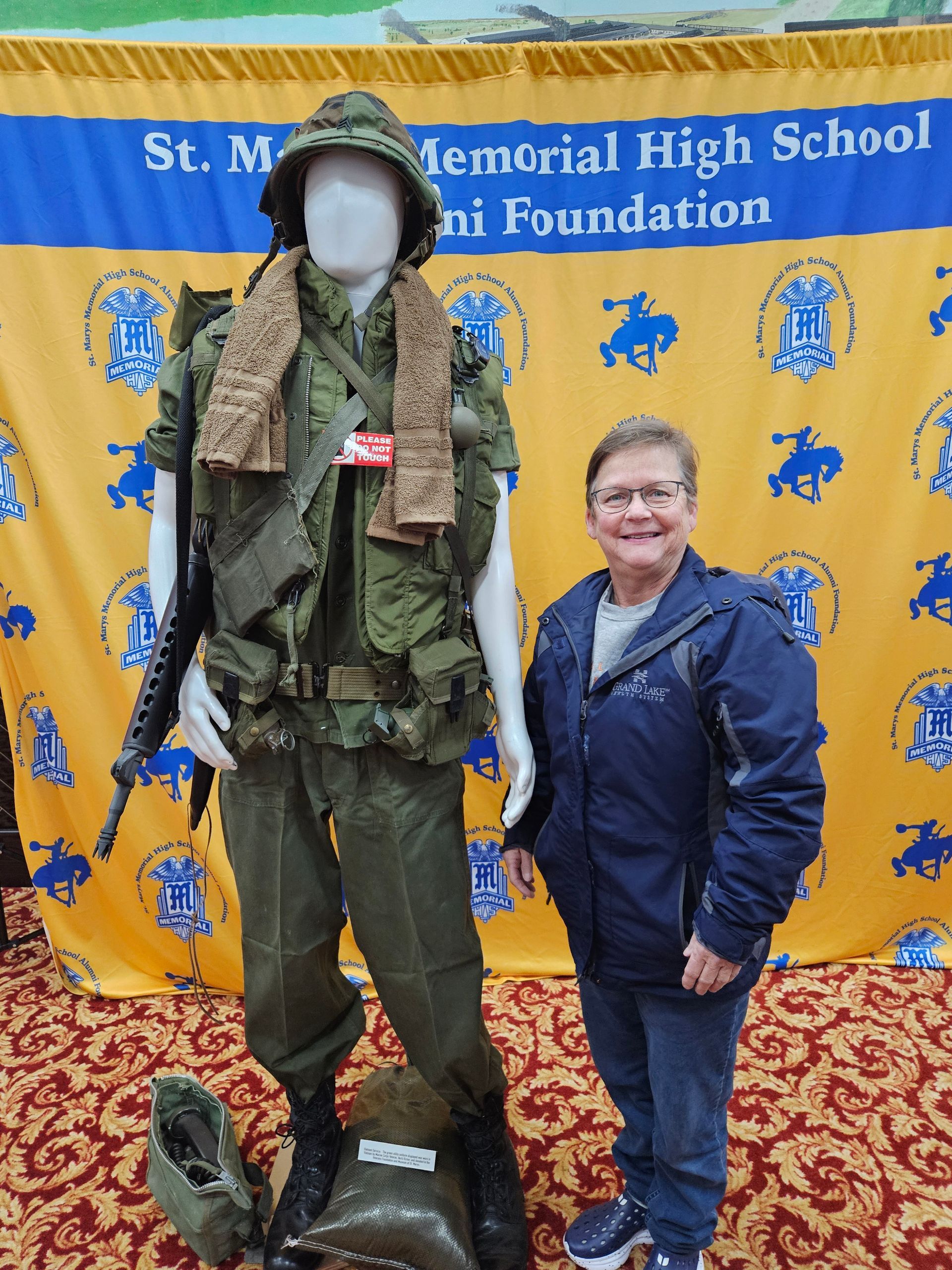 Woman stands next to a mannequin wearing a Vietnam War uniform; backdrop of school logo.
