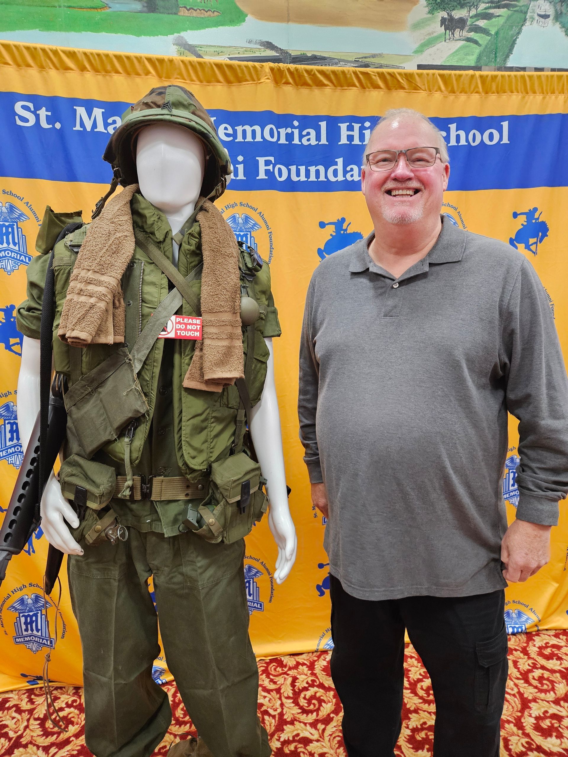 Man standing next to a mannequin wearing a Vietnam War uniform; both in front of a school banner.
