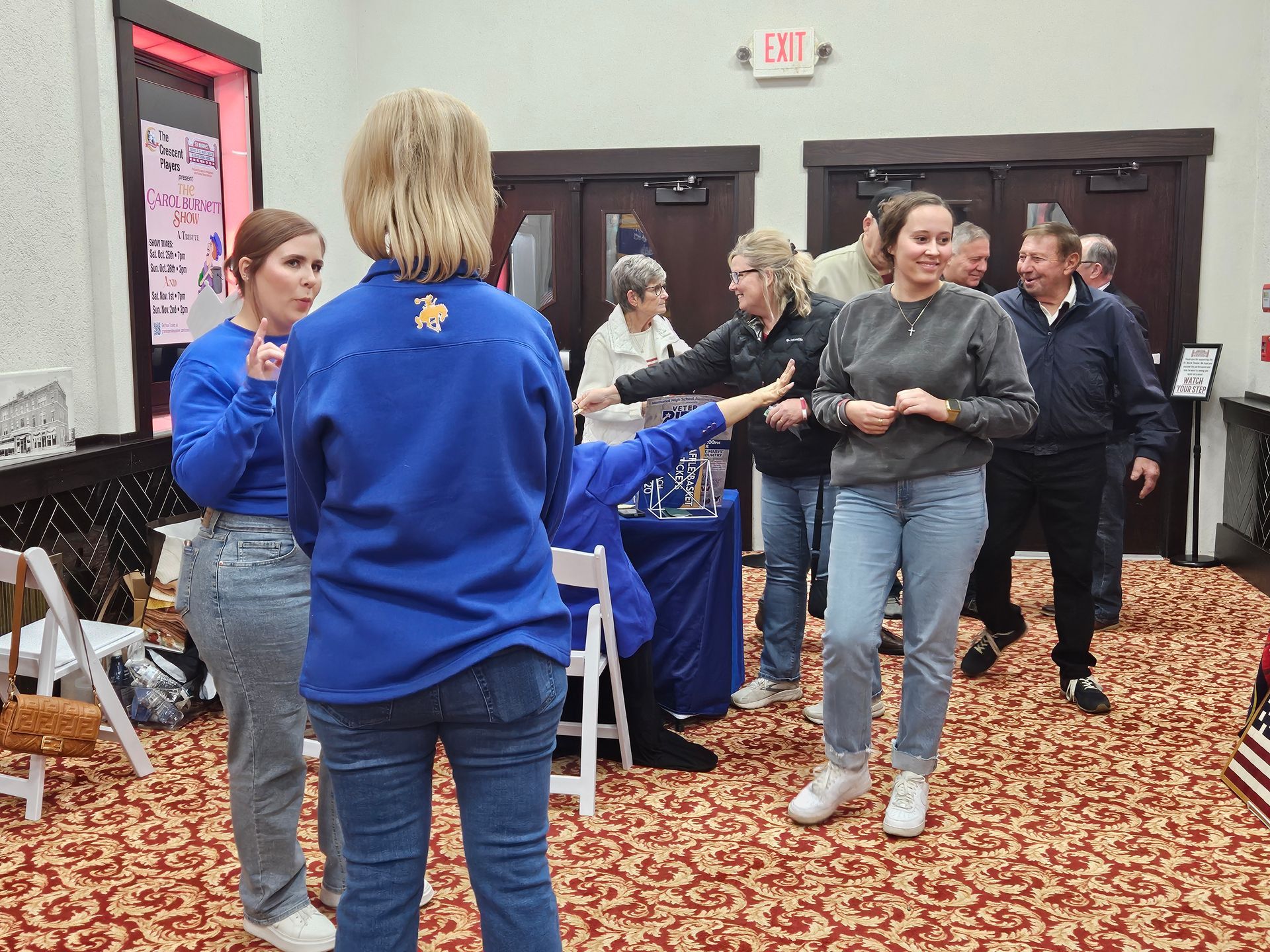 People in a room, some wearing blue, interacting at an event. Red carpet, tables, and an exit sign visible.