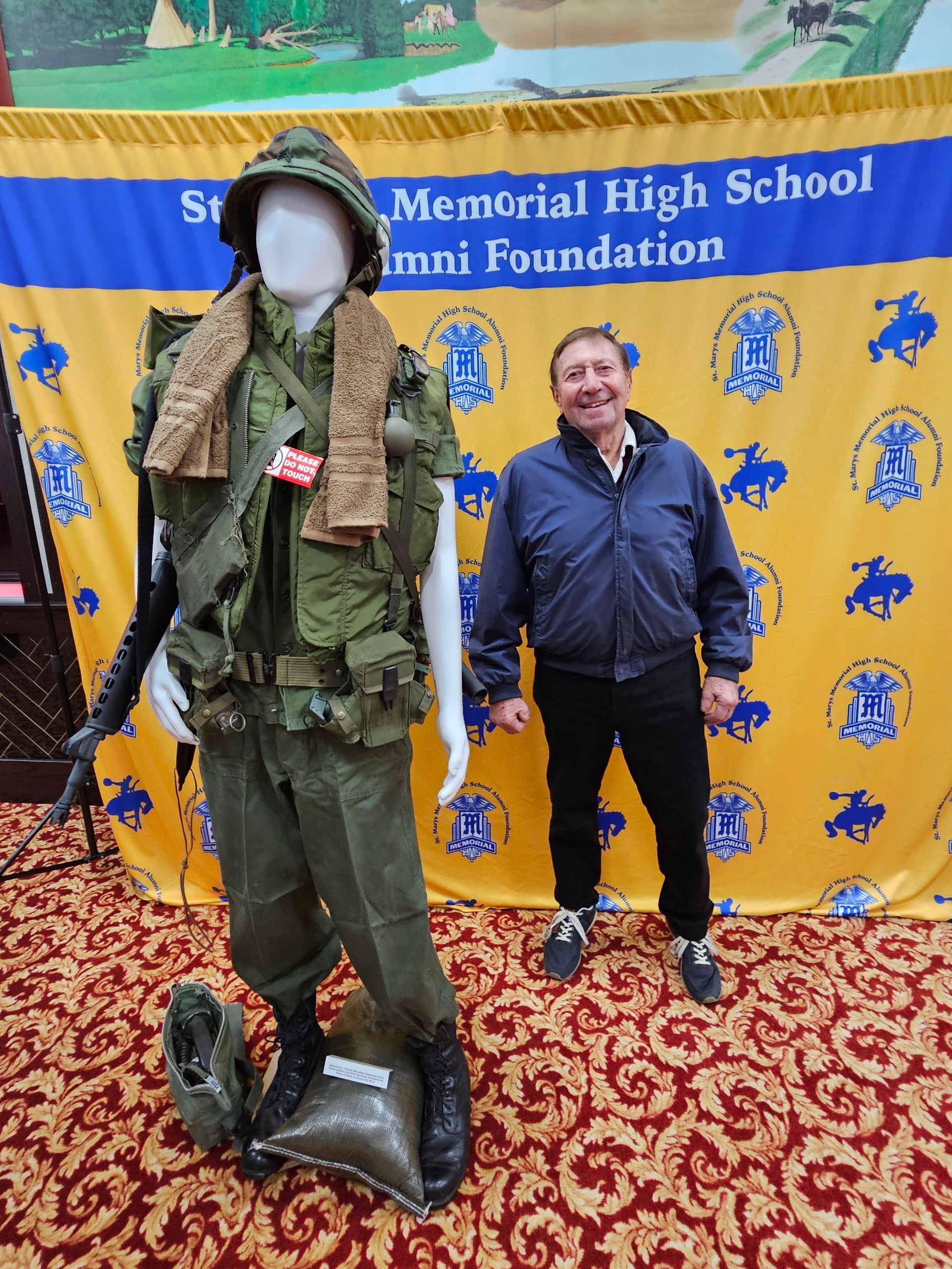 Man poses next to a mannequin dressed in a military uniform; backdrop features school logo.