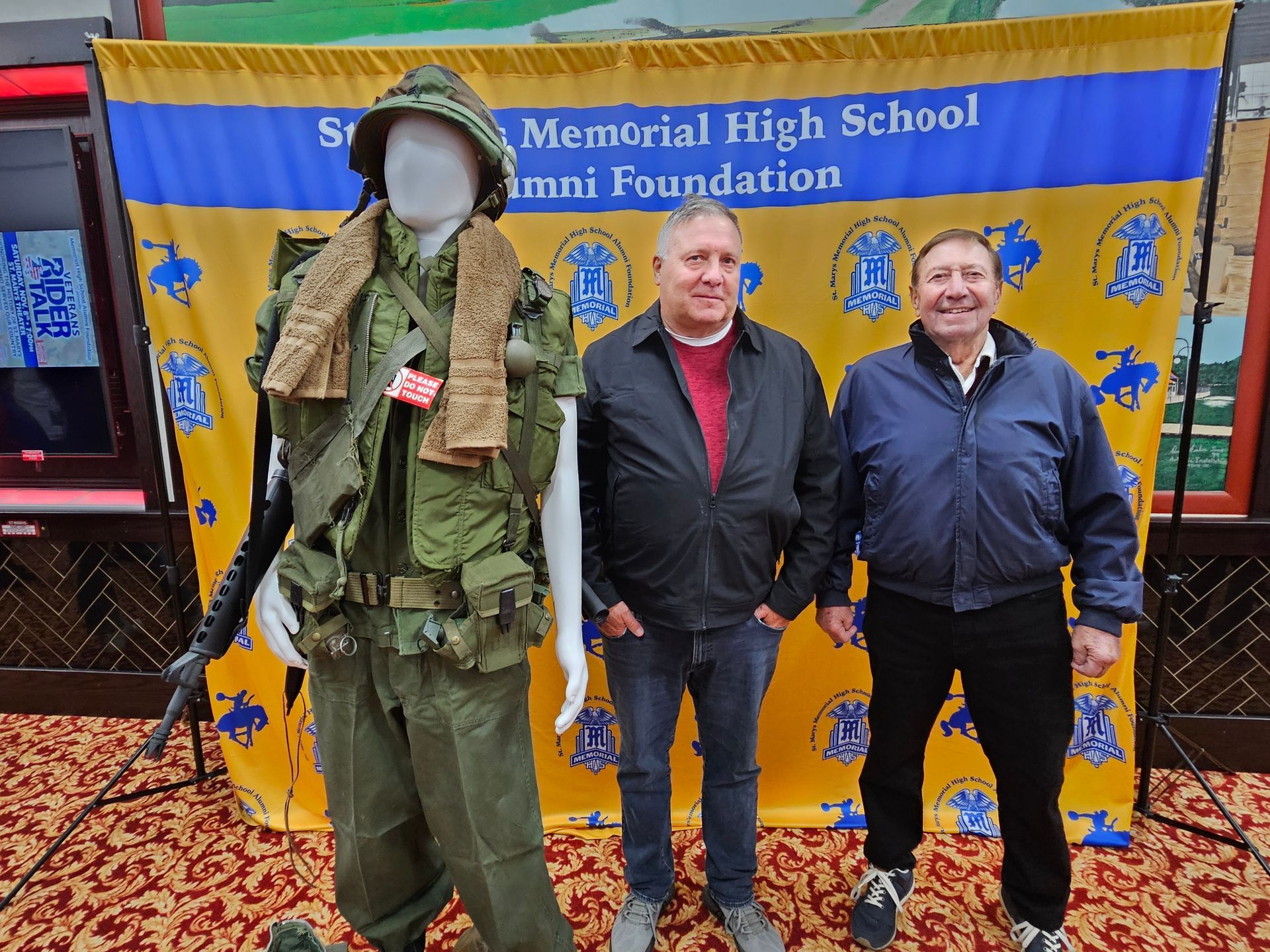 Two men stand by a mannequin in military uniform, in front of a banner for a high school alumni foundation.