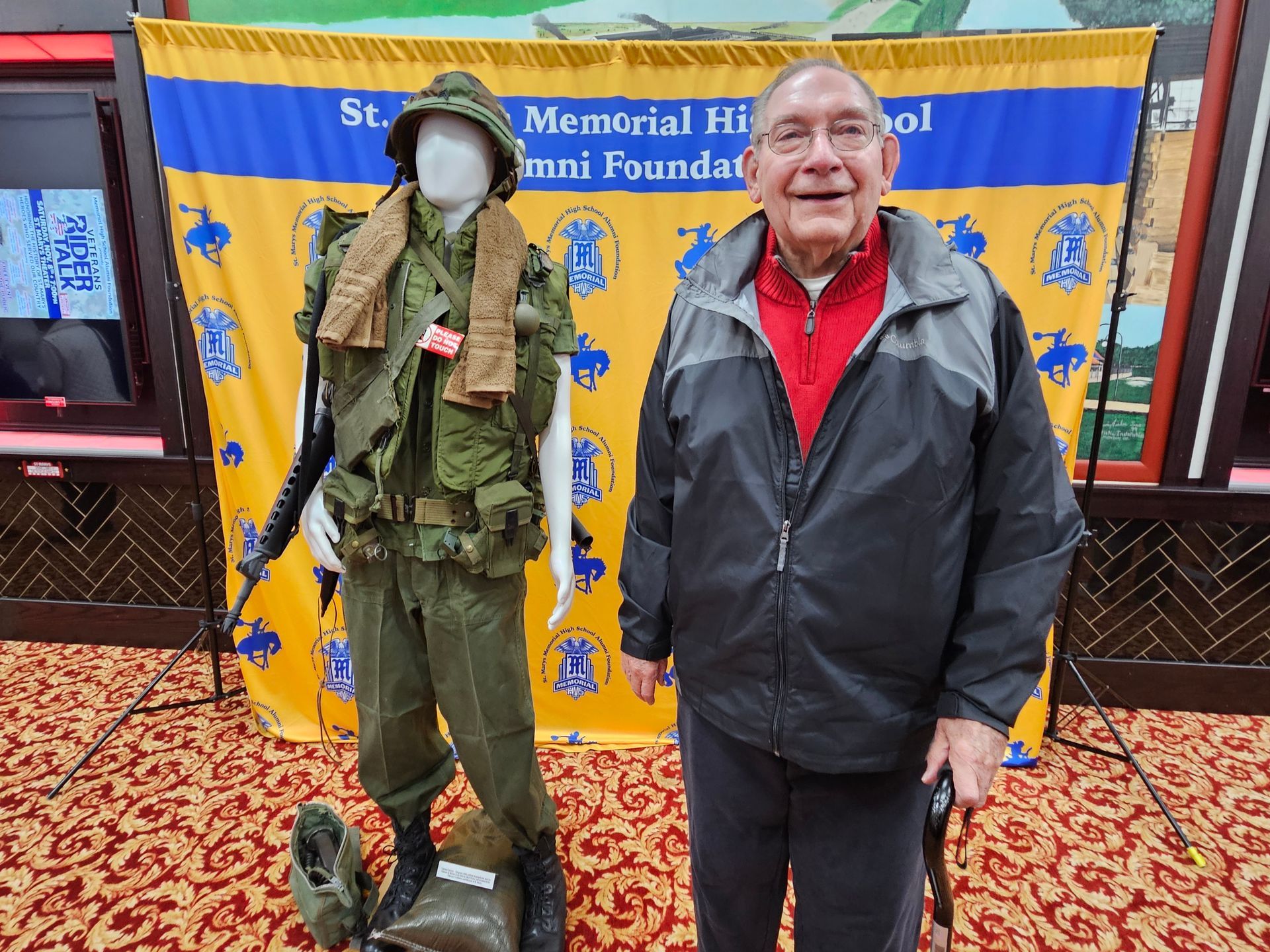 Man with cane stands next to a mannequin wearing a military uniform. Background is a school banner.
