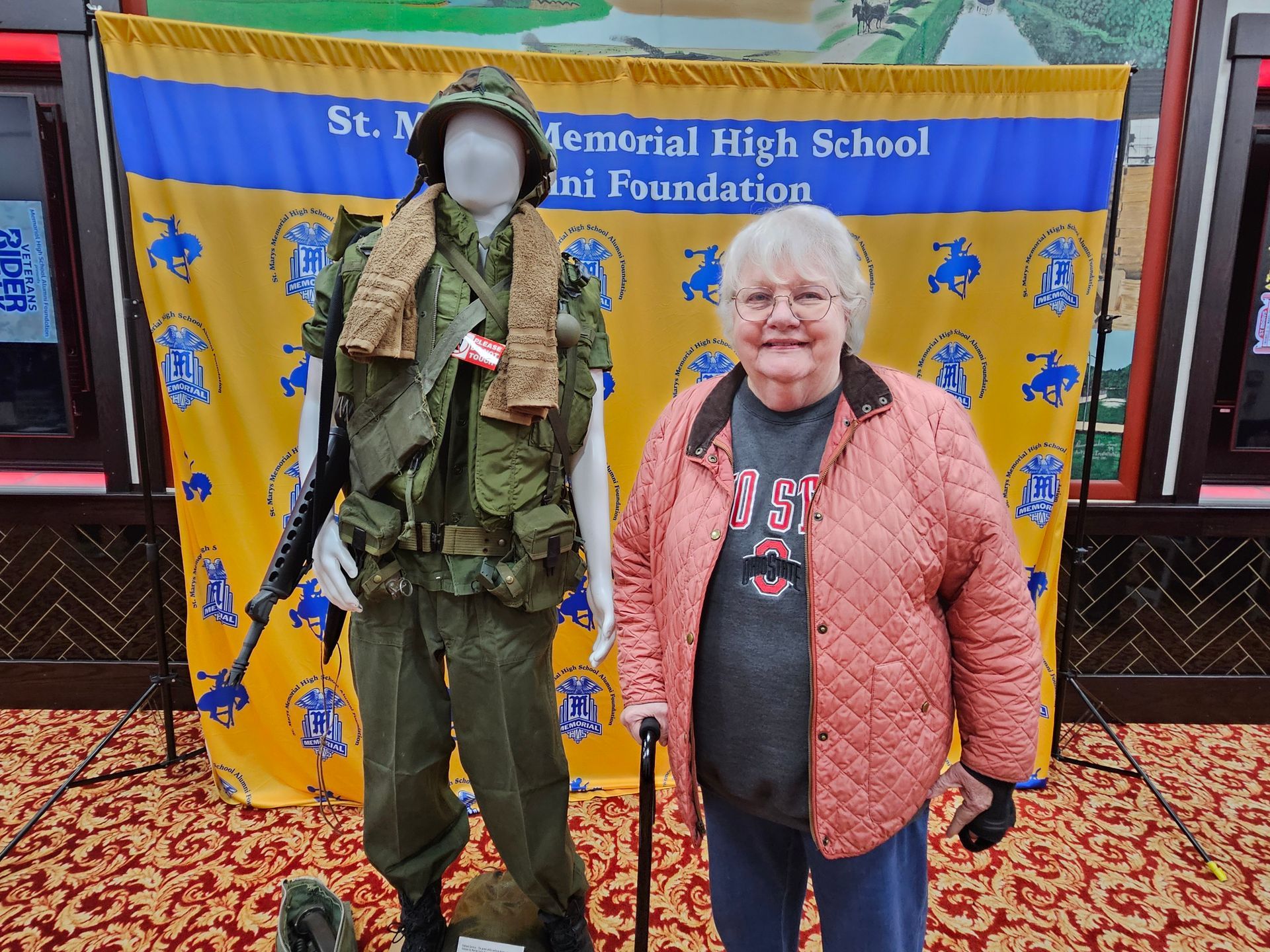 Woman poses next to a mannequin wearing military uniform. Background has school banner, mural, and carpet.